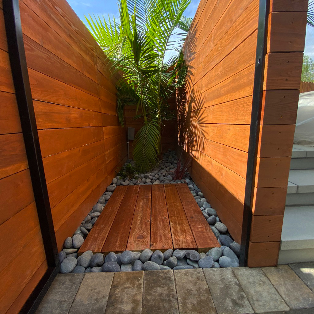 Narrow tropical garden passage with warm wooden walls, a palm, smooth river stones, and a wood walkway, creating a serene modern retreat.
