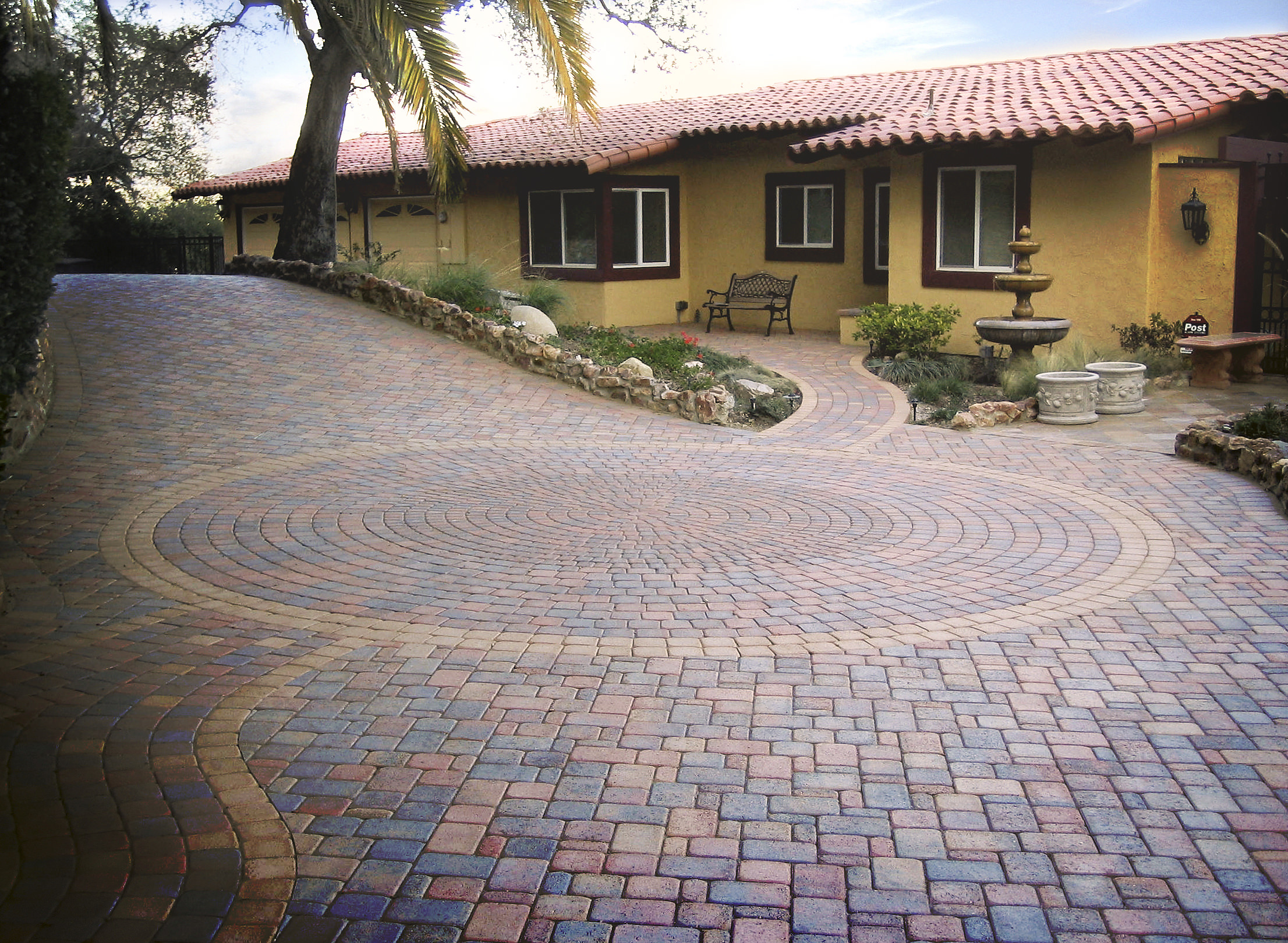 Mediterranean-style home with yellow stucco walls, red tile roof, lush landscaping, fountain, and a sweeping multicolored brick courtyard.