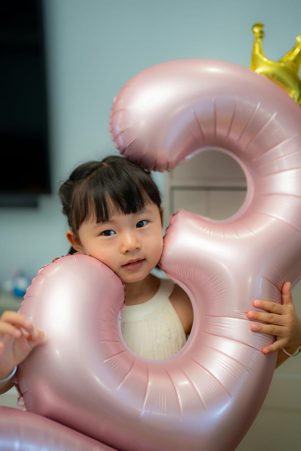 A young Asian girl with dark hair pulled back is holding two large, pink, heart-shaped balloons and a gold crown balloon behind her head. She is wearing a white sleeveless top. The background is softly blurred, showing a wall and a glimpse of a window or doorway.