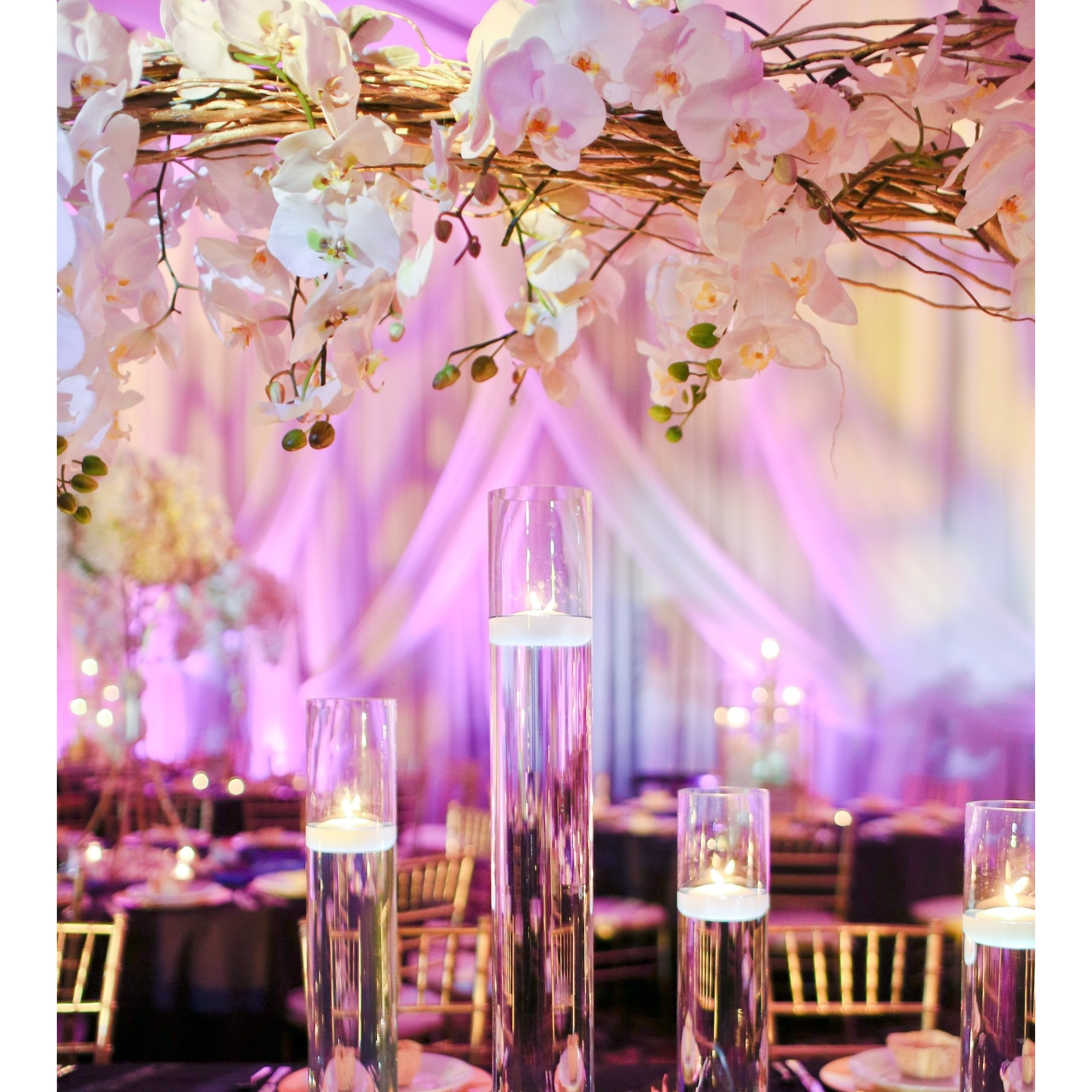 A stunningly romantic setting crafted by WS Event Production Inc., featuring elegant floral arrangements of white orchids cascading from golden branches. The soft pink and purple uplighting creates a dreamy and ethereal ambiance, perfectly complementing the floating candles in tall glass cylinders. The interplay of warm candlelight and vibrant colored lighting adds depth and dimension, creating an enchanting atmosphere ideal for a luxurious celebration.