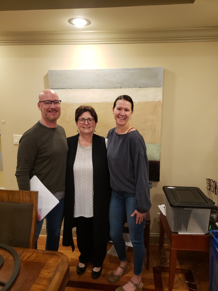 A man, a woman, and another woman stand smiling in front of a large abstract painting. The man on the left is wearing glasses and a dark long-sleeved shirt. The woman in the middle is wearing glasses, a black cardigan over a white and black polka-dot blouse, and black pants. The woman on the right is wearing a blue long-sleeved shirt and blue jeans.