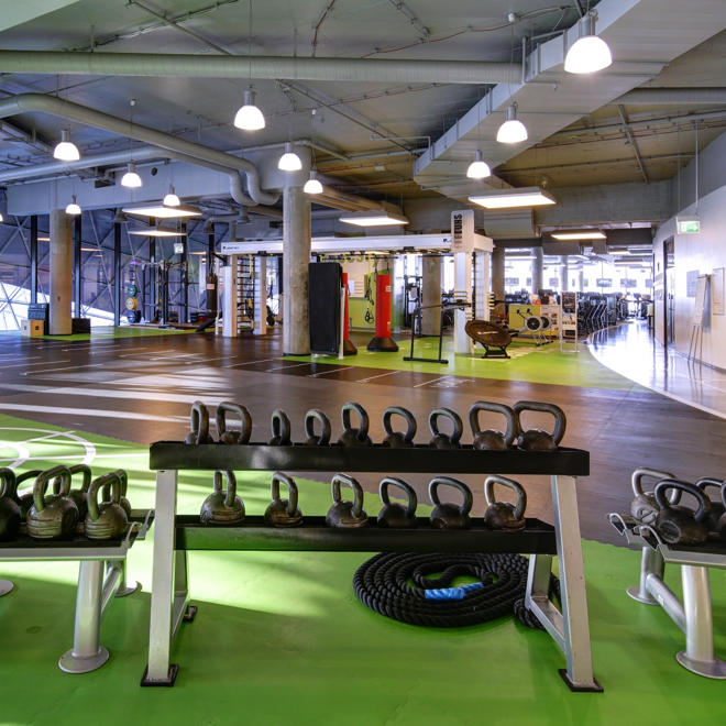 In einem Fitness-First-Standort, mit leuchtend grünen Matten, Regalen mit Kettlebells in allen Größen, einem aufgerollten Battle Rope und weiteren Trainingsgeräten im Hintergrund.