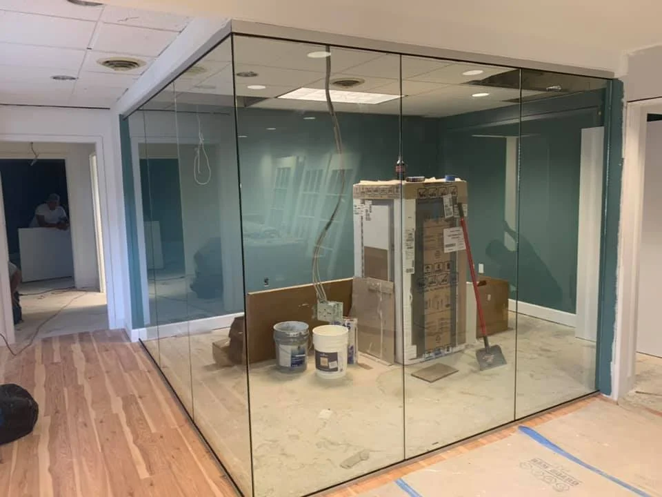 Construction or renovation work is underway in an office space, featuring a large, modern glass enclosure. Inside the enclosure, there are large cardboard boxes, a broom, and buckets of paint, suggesting ongoing interior work. Outside the enclosure, the floor is finished with polished wood. In the background, through an open doorway, a person in a white shirt is visible, along with some construction materials. The walls are painted a teal color. The ceiling has recessed lighting and exposed wires.