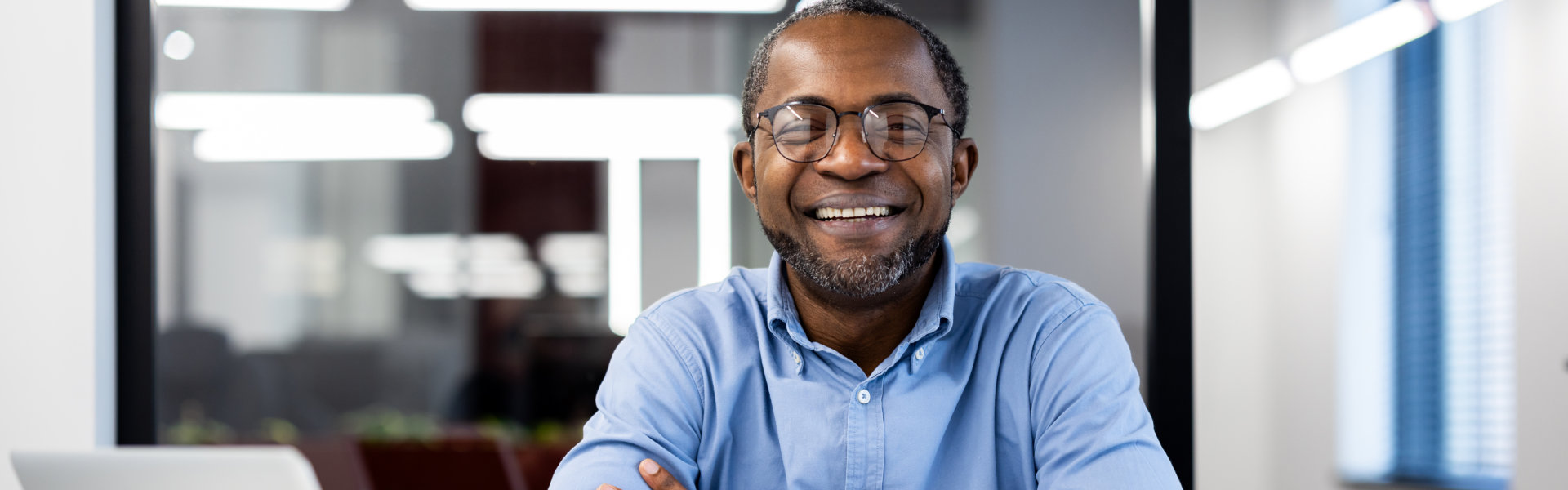 A middle-aged Black man with glasses and a beard, wearing a blue collared shirt, smiles broadly at t 