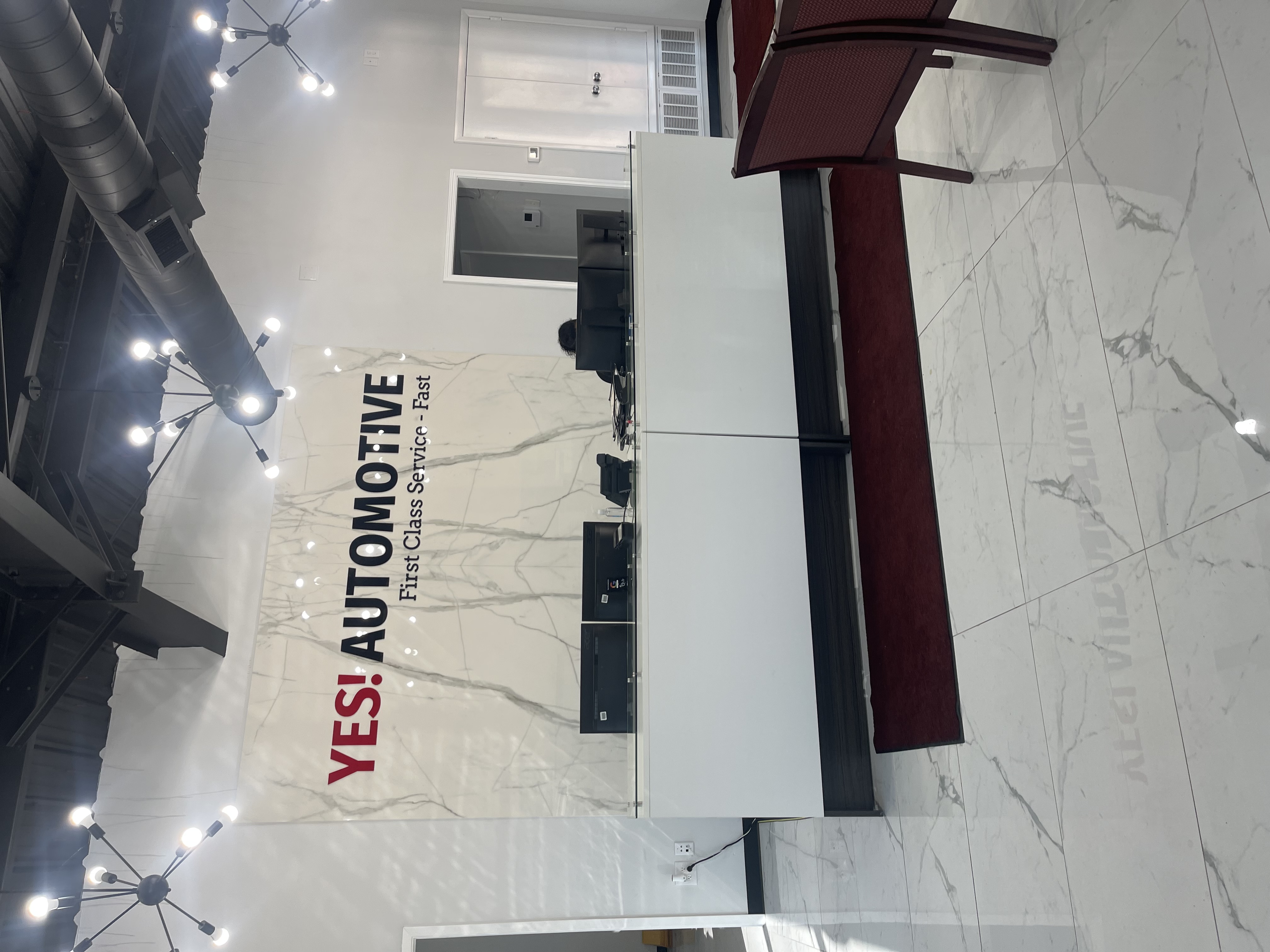 Front customer service counter inside the lobby of Yes! Automotive in Houston, Texas, featuring prominent wall signage reading "YES! AUTOMOTIVE - First Class Service - Fast" set against a marble-paneled backdrop.