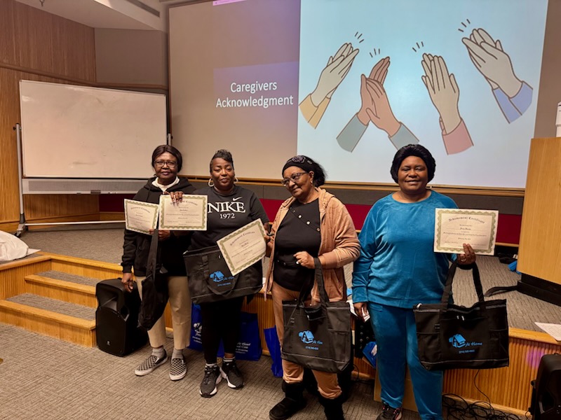 Four women are standing in a room with a projector screen behind them. They are holding certificates and tote bags. The projector screen has text that says "Caregivers Acknowledgment" and an illustration of hands clapping. The woman on the far left is wearing glasses and a black hoodie. The second woman from the left is wearing a Nike shirt. The third woman from the left is wearing a tan jacket and the woman on the far right is wearing a blue tracksuit. The certificates appear to be "Achievement Certificates". The tote bags are black with a blue logo and white text.