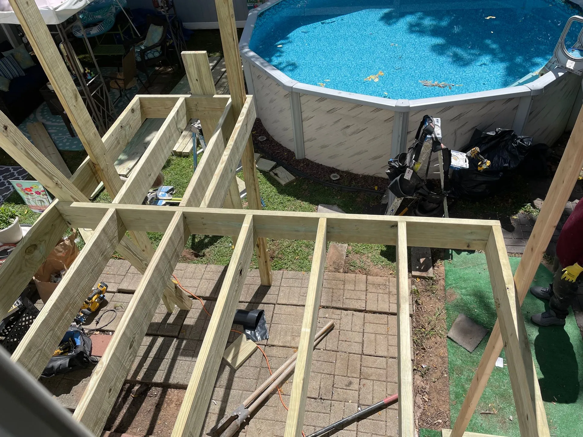 Wood framing for an elevated deck is under construction beside an above-ground swimming pool. Pressure-treated lumber supports future decking, demonstrating structural planning and residential outdoor improvement expertise.