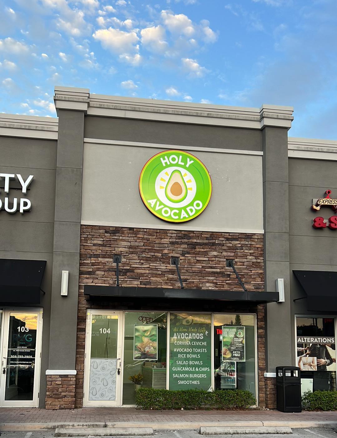 The front of the "Holy Avocado" business in Hallandale Beach, Florida, features a modern and clean design. The facade is gray with a section adorned with bricks in brown and cream tones, giving it an attractive texture and contrast. Centered above the entrance is a large circular sign with the business's logo: the name "Holy Avocado" in white letters on a green background, with an image of an avocado at the center, emphasizing the restaurant's theme.

There are two large windows allowing a peek inside, over which there are vinyl decals showcasing the products they offer, such as "Avocado Toasts," "Rice Bowls," "Salad Bowls," "Guacamole & Chips," "Salmon Burgers," and "Smoothies." The "Corvina Ceviche" is also highlighted, suggesting the freshness of the ingredients used. The unit number is 104, and right next to it, at 103, there appears to be another business named "QUALITY CUP," though it is not the focus of the image.

The sidewalk in front of the business is clear with several parking spaces available. The lighting appears to be ample, with at least one light fixture visible above the door, likely providing excellent visibility during nighttime hours. The clear sky with scattered clouds suggests the photo was taken on a clear day. Overall, the establishment conveys a sense of being a clean and welcoming place that specializes in healthy dishes with avocado as a key ingredient.
