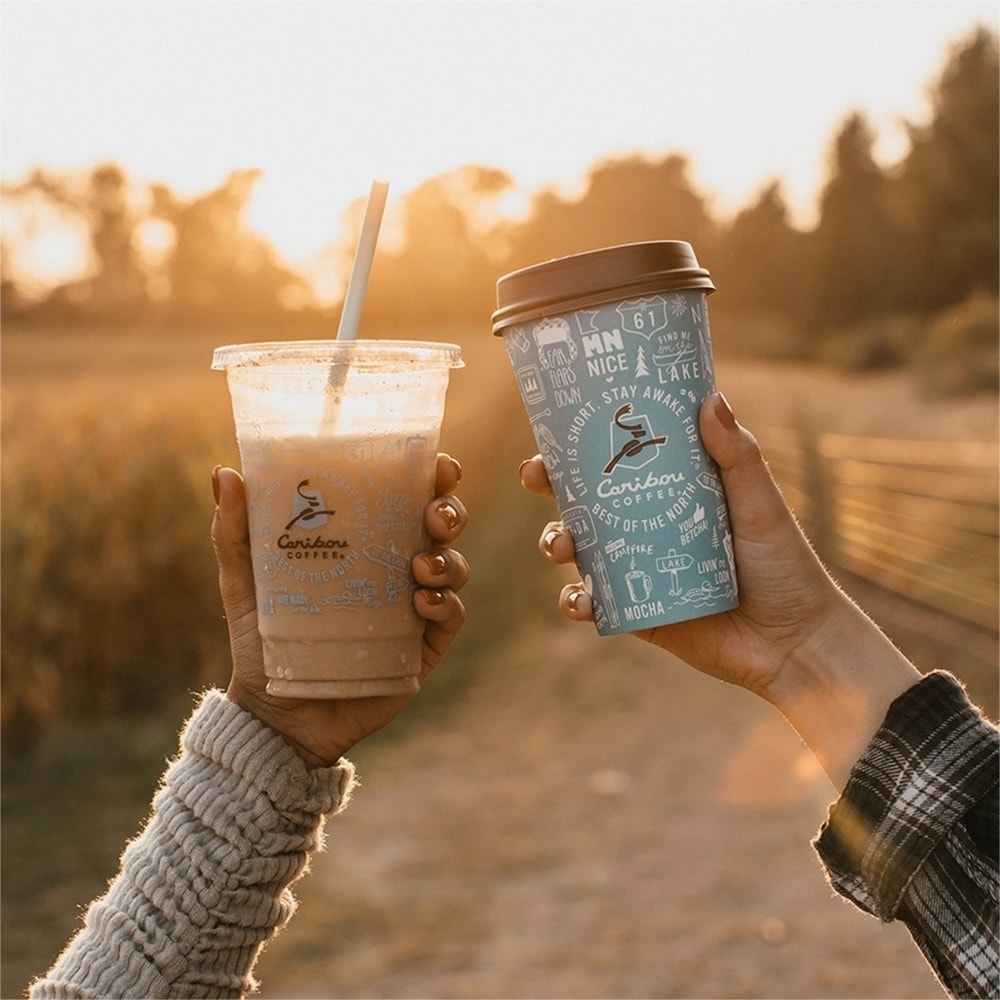 Une main tenant un café glace dans un gobelet plastique Caribou Coffee et une autre main tenant un café chaud dans un gobelet Caribou Coffee classique