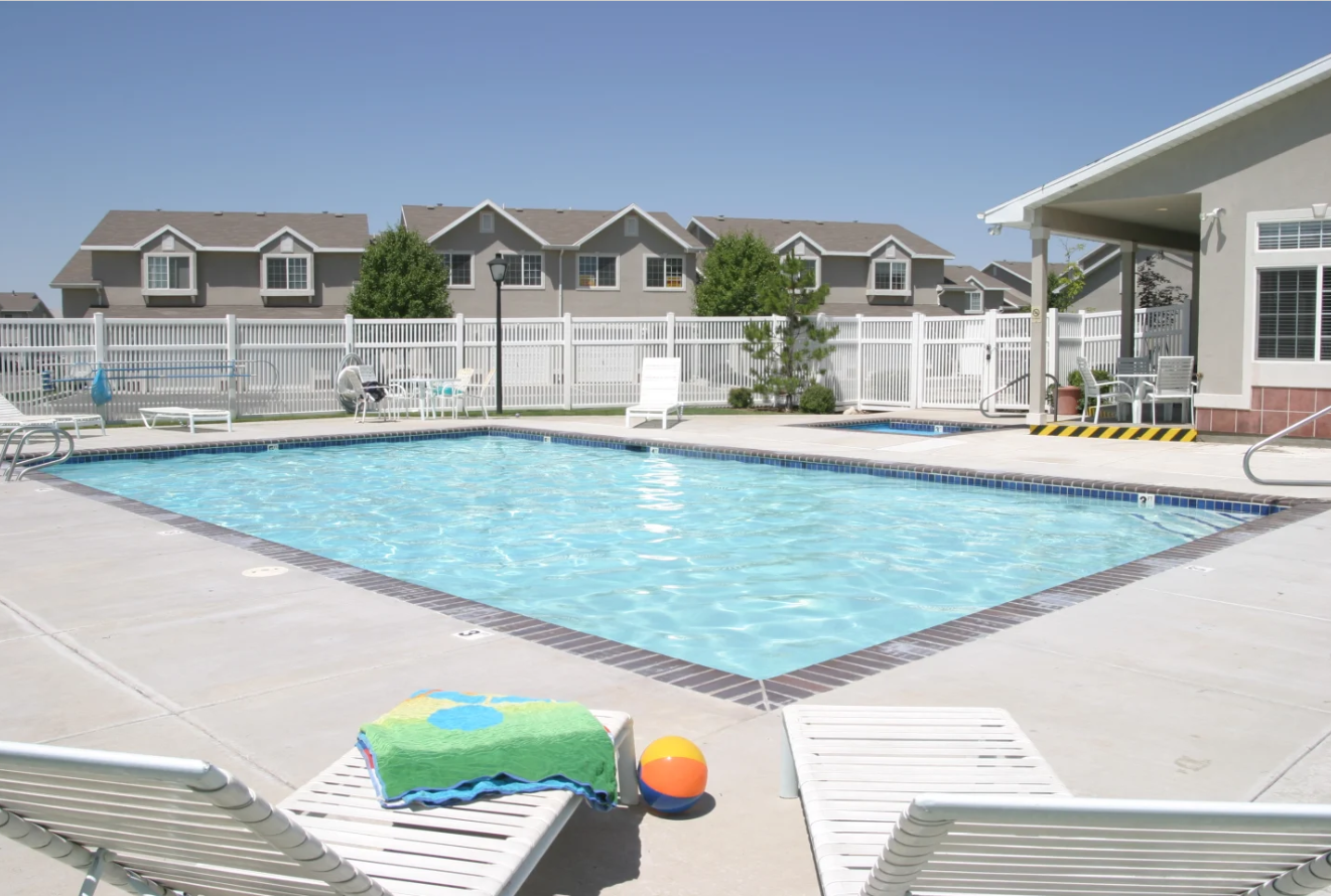 View of the blue swimming pool with a white fence, white chaise lounge chairs with a colorful towel on one and a multicolored beach ball in between, and resident building to the right