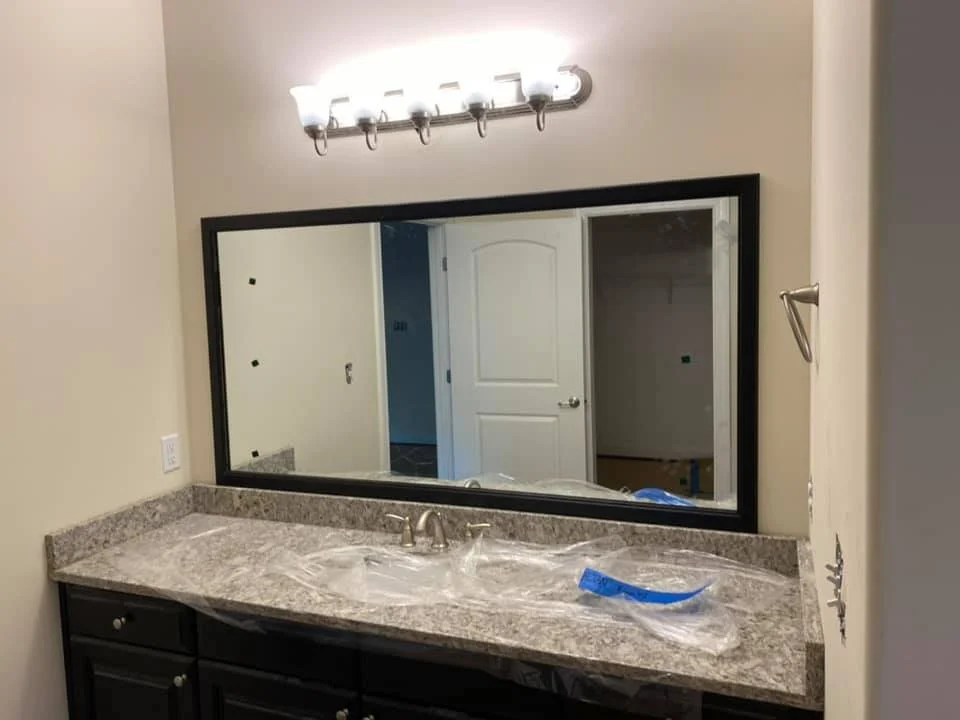 A wide framed mirror above a granite countertop in a bathroom with plastic sheeting covering the countertop and sink area. A light fixture with four bulbs is mounted above the mirror, and a towel bar is visible on the right wall.