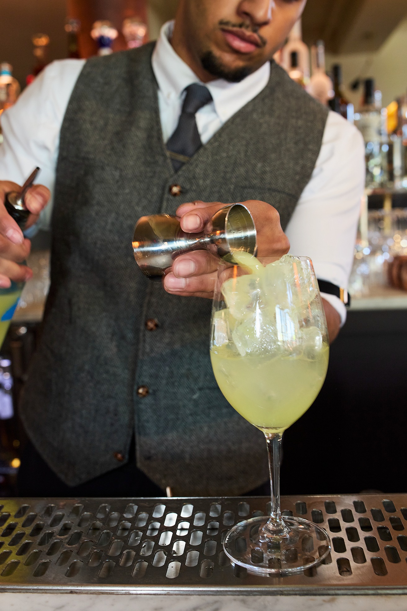 A bartender in a grey vest and tie carefully pours a pale yellow cocktail from a silver jigger into a glass filled with ice. The action takes place over a stainless steel bar rail.
