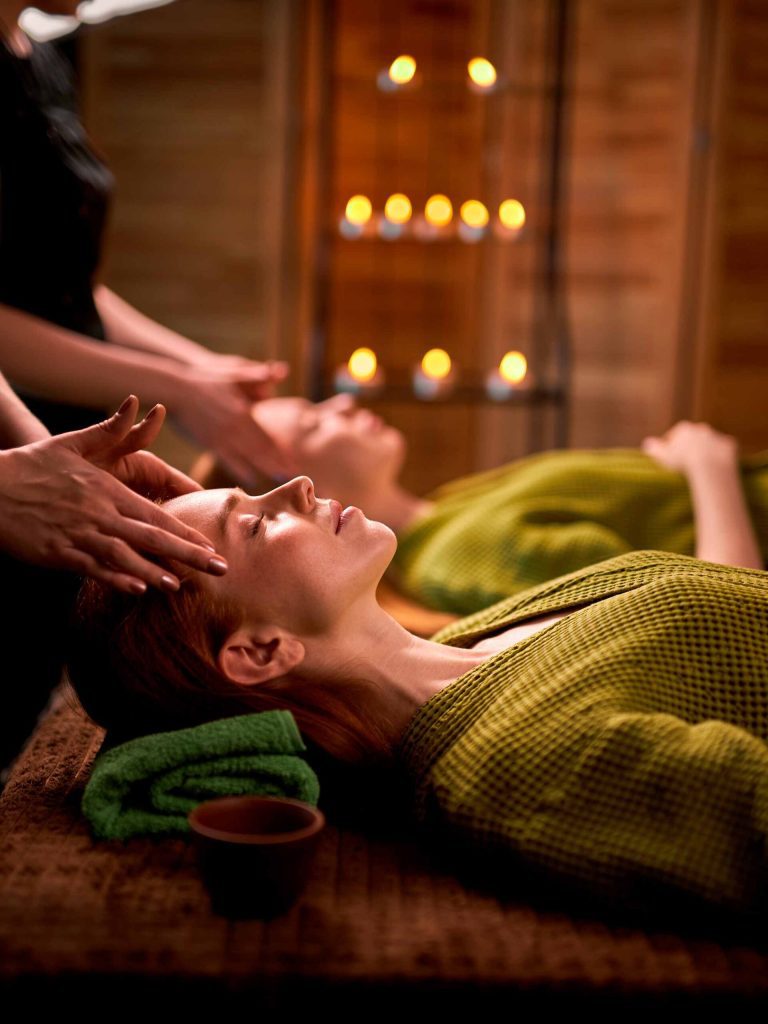 Two women are receiving massages in a dimly lit spa, with candles flickering in the background. The woman in the foreground has red hair and is wearing a green robe. Her eyes are closed, and her face is tilted upwards as if enjoying the experience. A massage therapist's hands are gently massaging her temples. Another woman in the background is also lying down, presumably also receiving a massage. The spa's interior features wooden walls and soft lighting, creating a relaxing atmosphere.