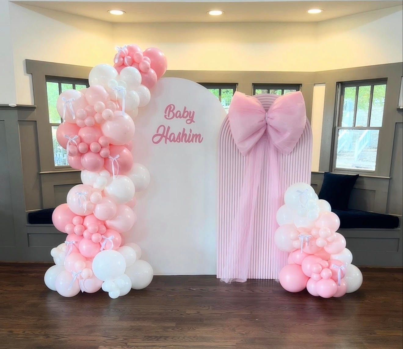 Elegant baby shower backdrop featuring soft pink and white balloon garlands, arched panels, pleated fabric with bow accent, personalized Baby Hashim sign, creating a charming, celebratory indoor event display setting.
