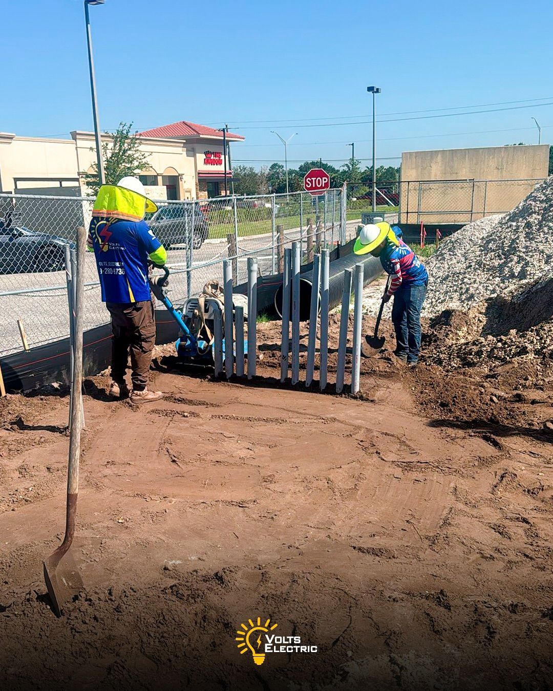 Two electricians working outdoors at a construction site installing underground electrical conduits, using tools to compact soil and position pipes near a fenced area beside a commercial roadway.