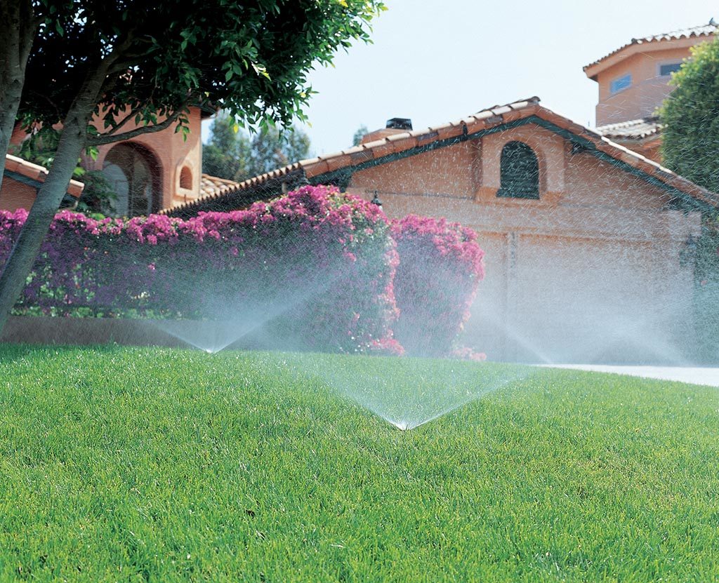 A residential lawn sprinkler system watering a lush green yard and landscaped beds in front of a house.