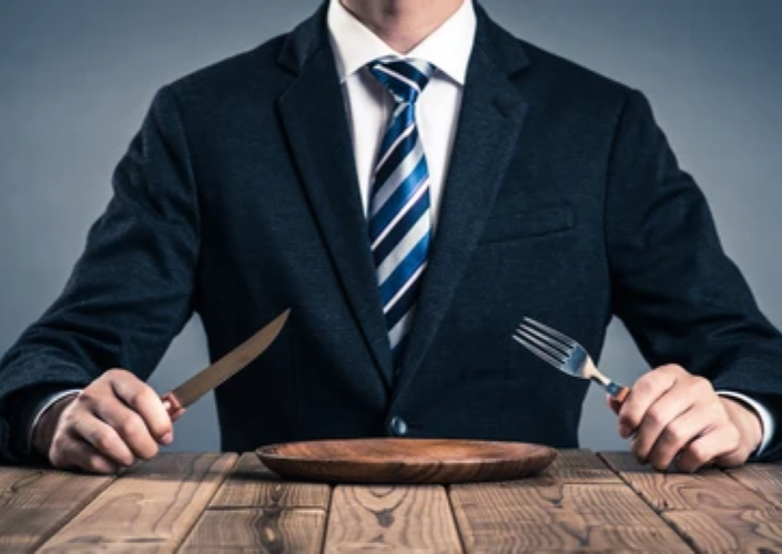 Man in a suit sitting at a table in front of an empty plate and holding a knife and fork.