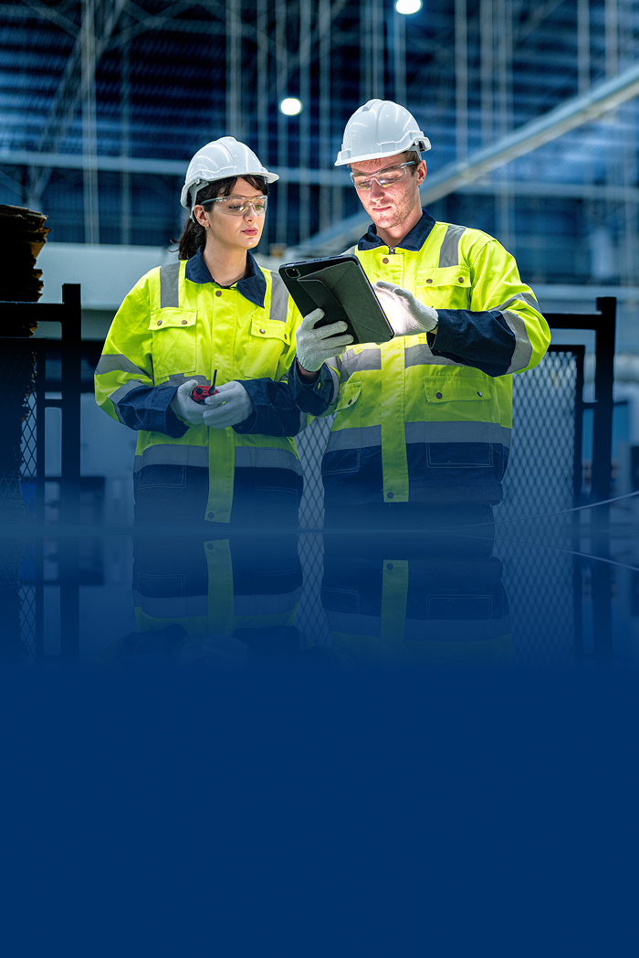 Two engineers in a factory wearing hard hats and work clothes and looking at a tablet.