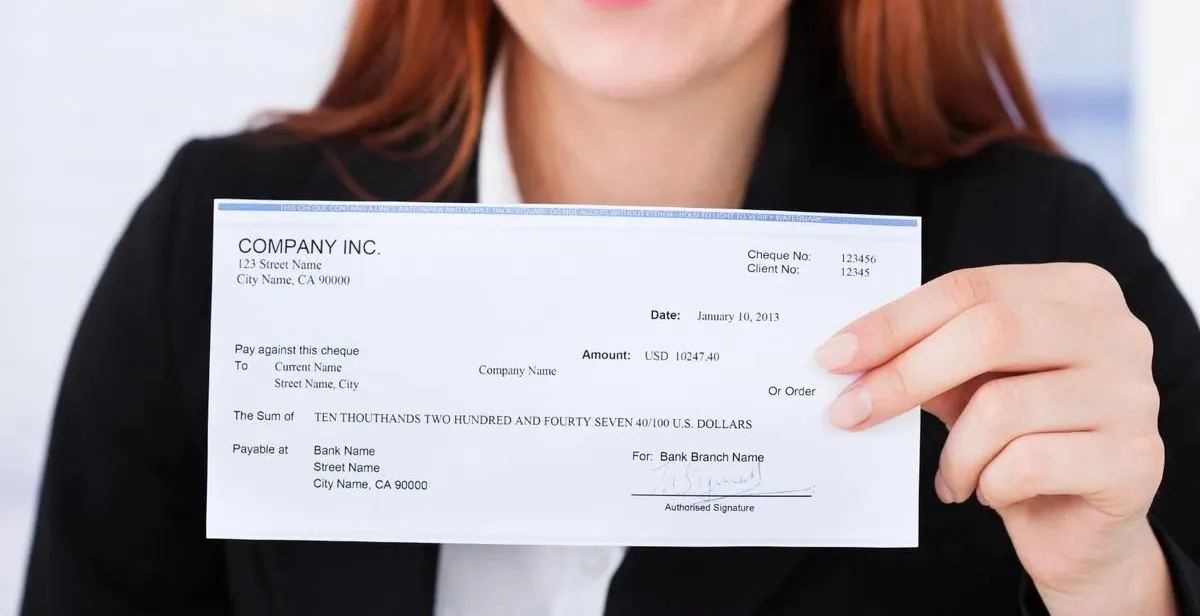 A woman in a black blazer holds up a check from "COMPANY INC.". The check is dated January 10, 2013, and is for the amount of USD 10247.40, written out as "TEN THOUSAND TWO HUNDRED AND FOURTY SEVEN 40/100 U.S. DOLLARS".