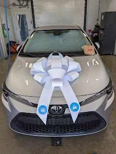 A silver Toyota sedan with a large white bow on its hood sits in what appears to be a showroom or service bay, ready for delivery or display. The bow has blue circular logos with white icons on its streamers. A small gift bag rests on the windshield.