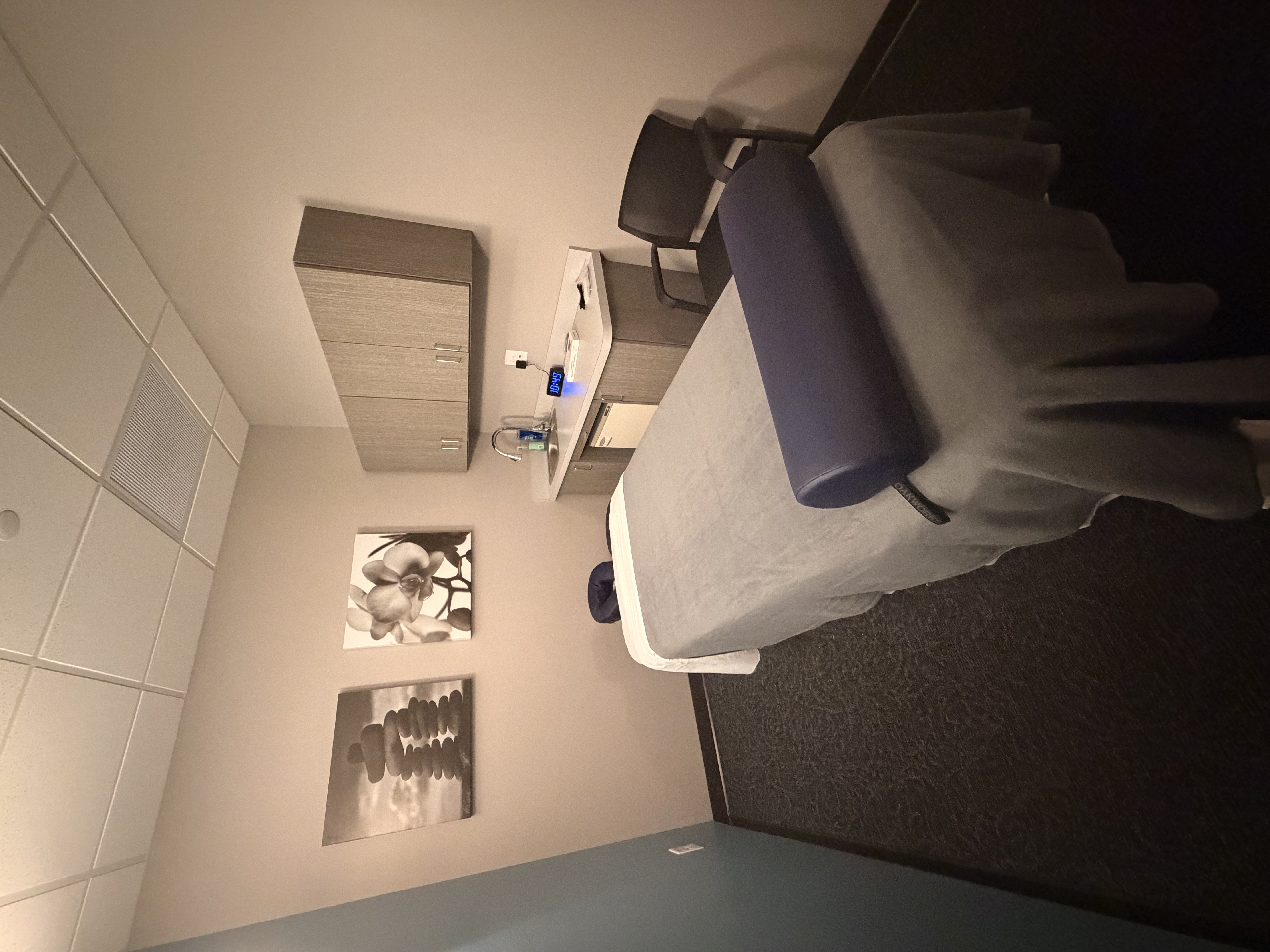 Massage room with treatment table in gray linens and navy blue face cradle cover. Gray wood-grain cabinet with sink. Digital clock on counter. Black and white floral canvas art on wall. Soft ambient lighting and neutral color palette.