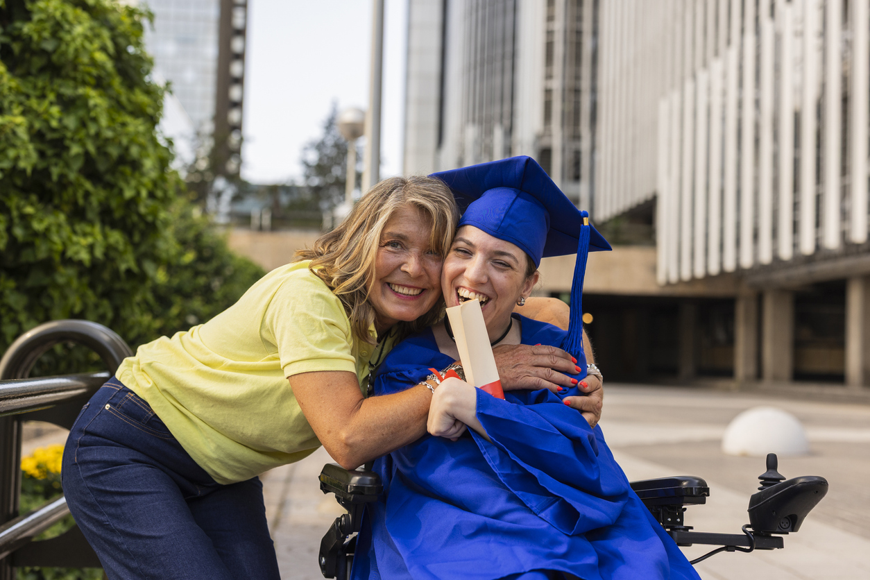 A woman in a wheelchair wearing a blue graduation gown and cap smiling, hugs another woman in a yellow top outdoors. They both appear joyful and celebratory.