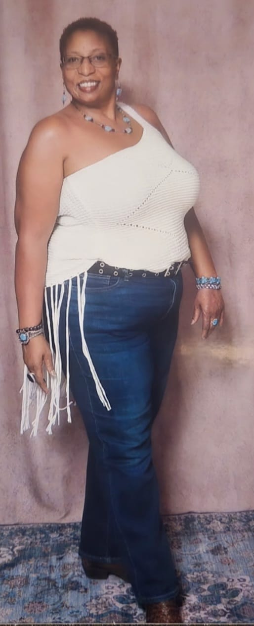 A full-body portrait of a smiling Black woman wearing glasses, a white one-shoulder top with fringe, blue jeans, and jewelry. She is standing in front of a pinkish-brown textured background and a patterned rug. The woman is wearing a necklace with blue stones, blue earrings, a bracelet on her left wrist, and rings on her fingers. Her top has a crocheted texture and white fringe hanging from the hem. She is also wearing a black belt with silver grommets and dark blue flared jeans. Her shoes appear to be brown and possibly have a patterned texture.
