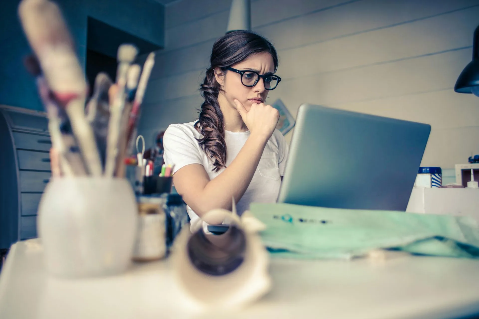A young woman with glasses is sitting at a desk, looking intently at a laptop screen. Her hand is resting on her chin in a thoughtful pose. In the foreground, a vase filled with paintbrushes and a roll of paper are visible. The scene suggests a creative workspace.