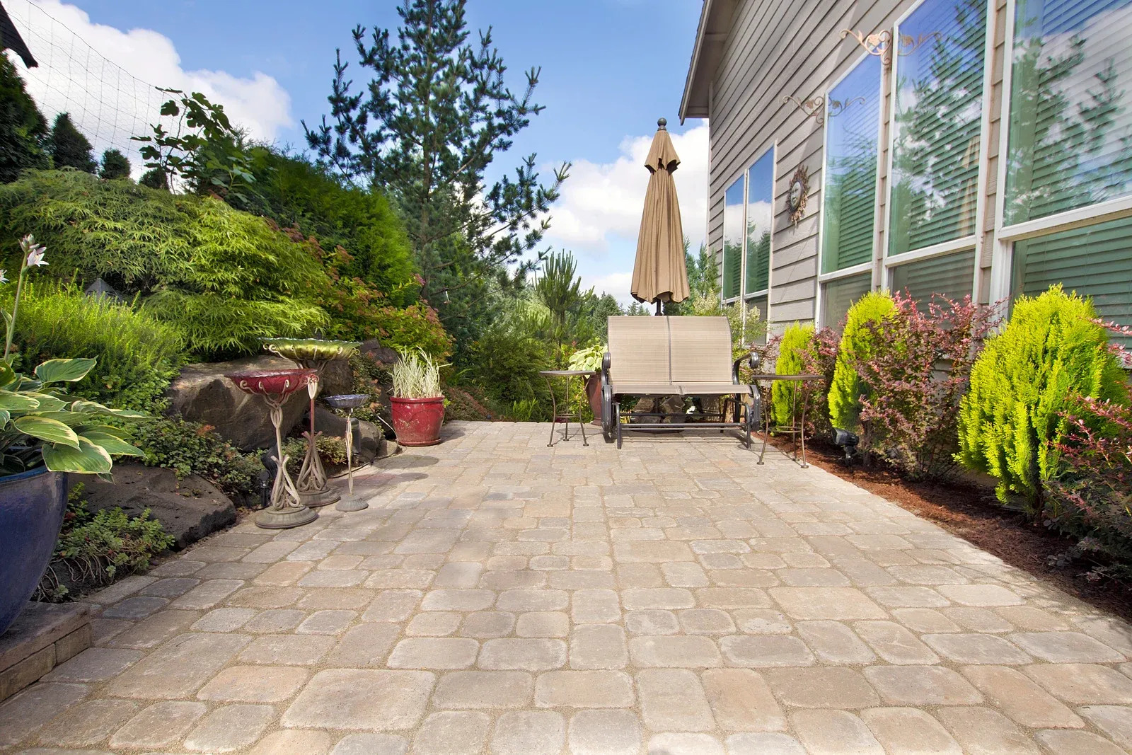 A spacious paver patio extends from a house, featuring outdoor furniture with a closed umbrella, surrounded by diverse landscaping, including a lush green hillside and various plants.