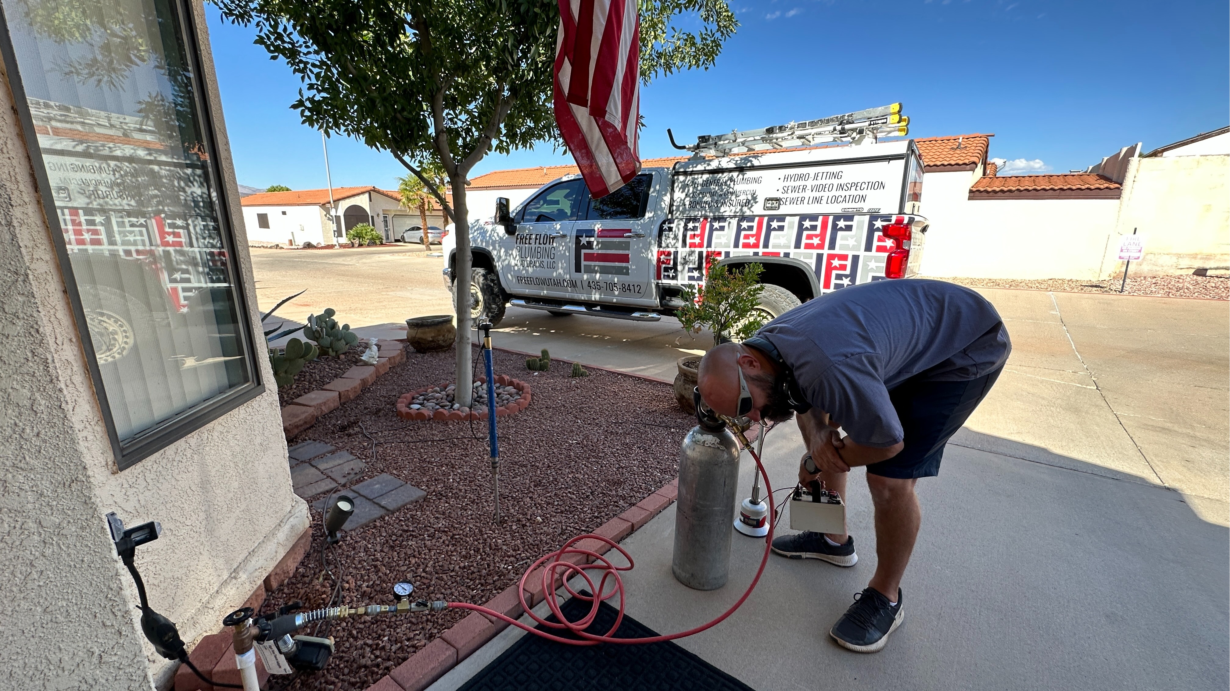 A Free Flow Plumbing and Drains technician is shown performing a residential service call in a St. George neighborhood. The company’s wrapped service truck is parked in the background, displaying the recognizable logo and service list. The technician is actively using plumbing diagnostic equipment, highlighting the company’s hands-on expertise and commitment to reliable, professional service at home visits.