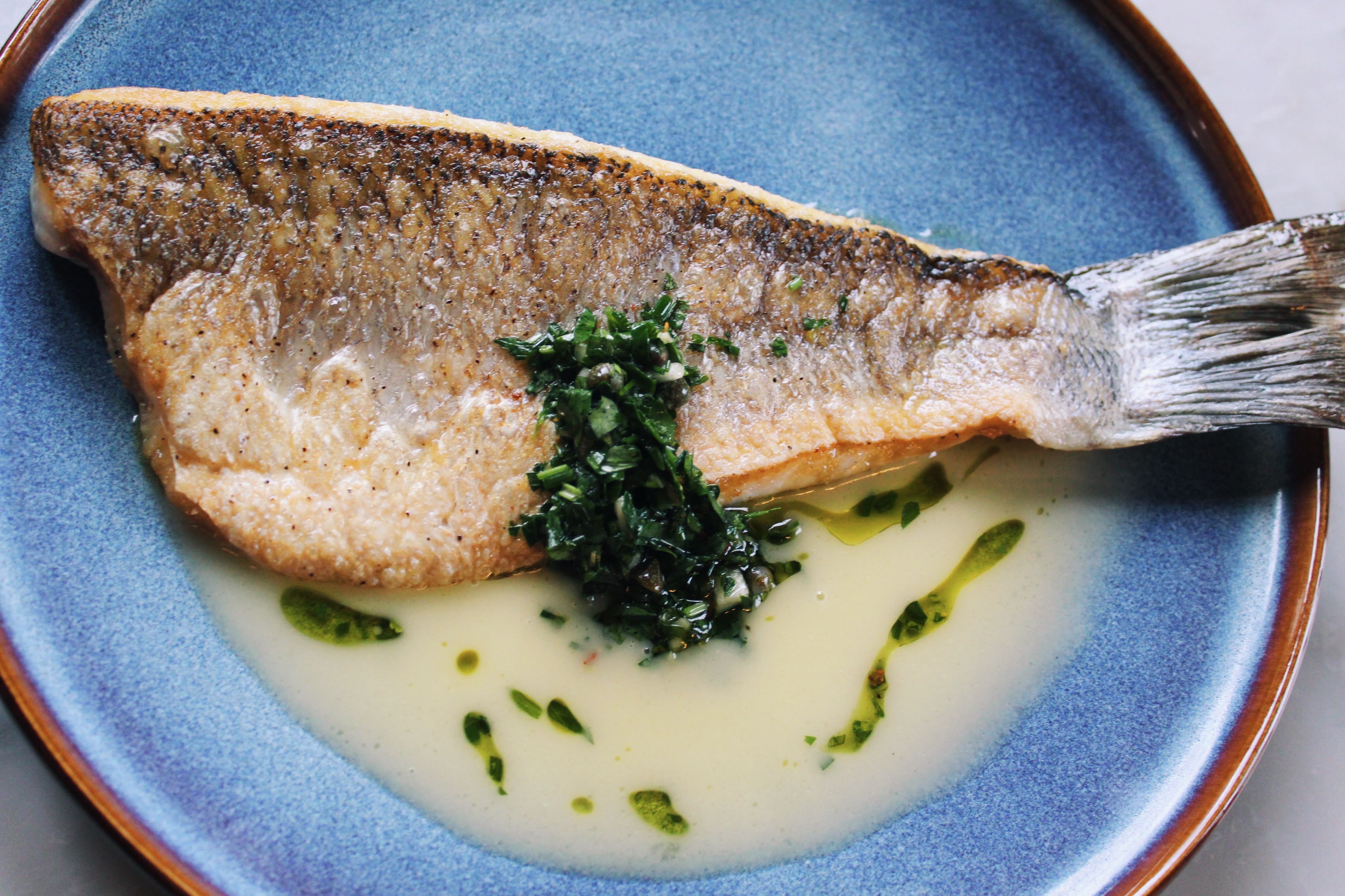 Our Crispy Skin Branzini — golden, crackling skin over delicate white fish, resting in silky lemon butter with a spoonful of fresh salsa verde and herb oil.
