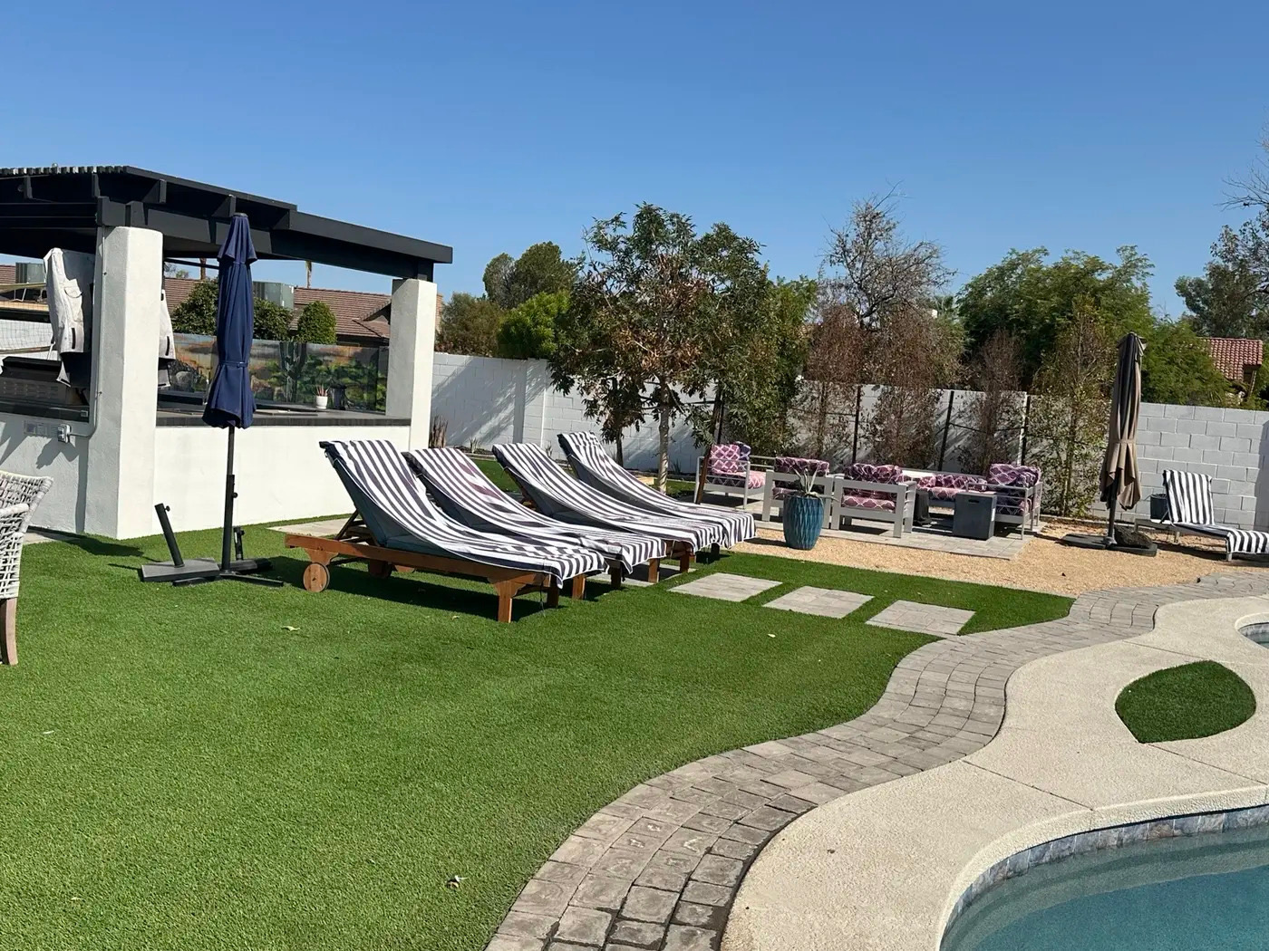 A sunny backyard oasis with a swimming pool, artificial turf, and lounge areas. Several lounge chairs with striped cushions are arranged on the turf, facing a covered outdoor kitchen/bar area. Further back, a seating area with a fire pit is set against a brick wall, surrounded by trees and shrubs. A paved pathway leads around the pool, which is partially visible in the foreground.