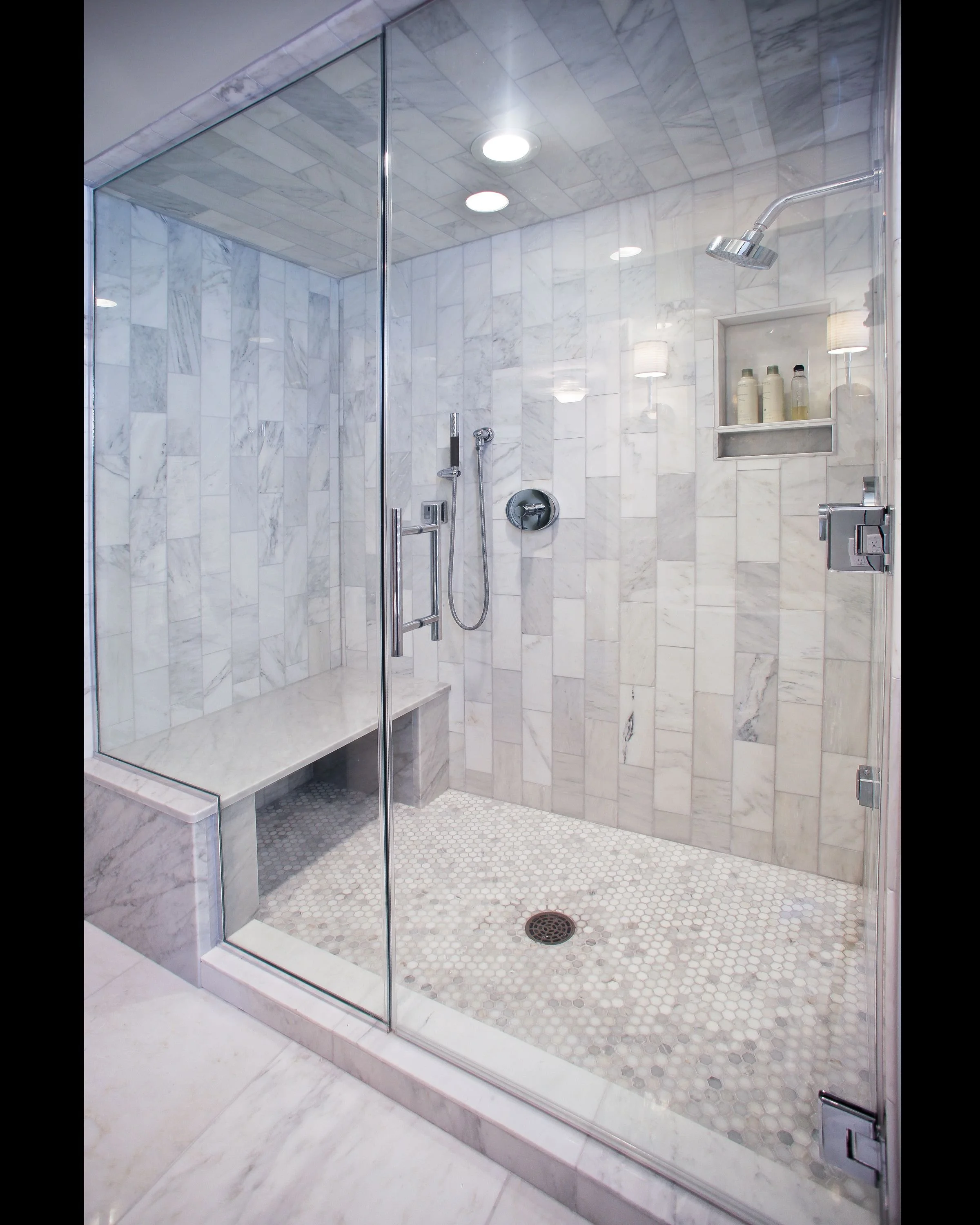 A modern and luxurious walk-in shower with marble tile walls and floor, a built-in bench, and a glass enclosure. The shower features a rainfall showerhead, a handheld shower, and a recessed niche for toiletries.