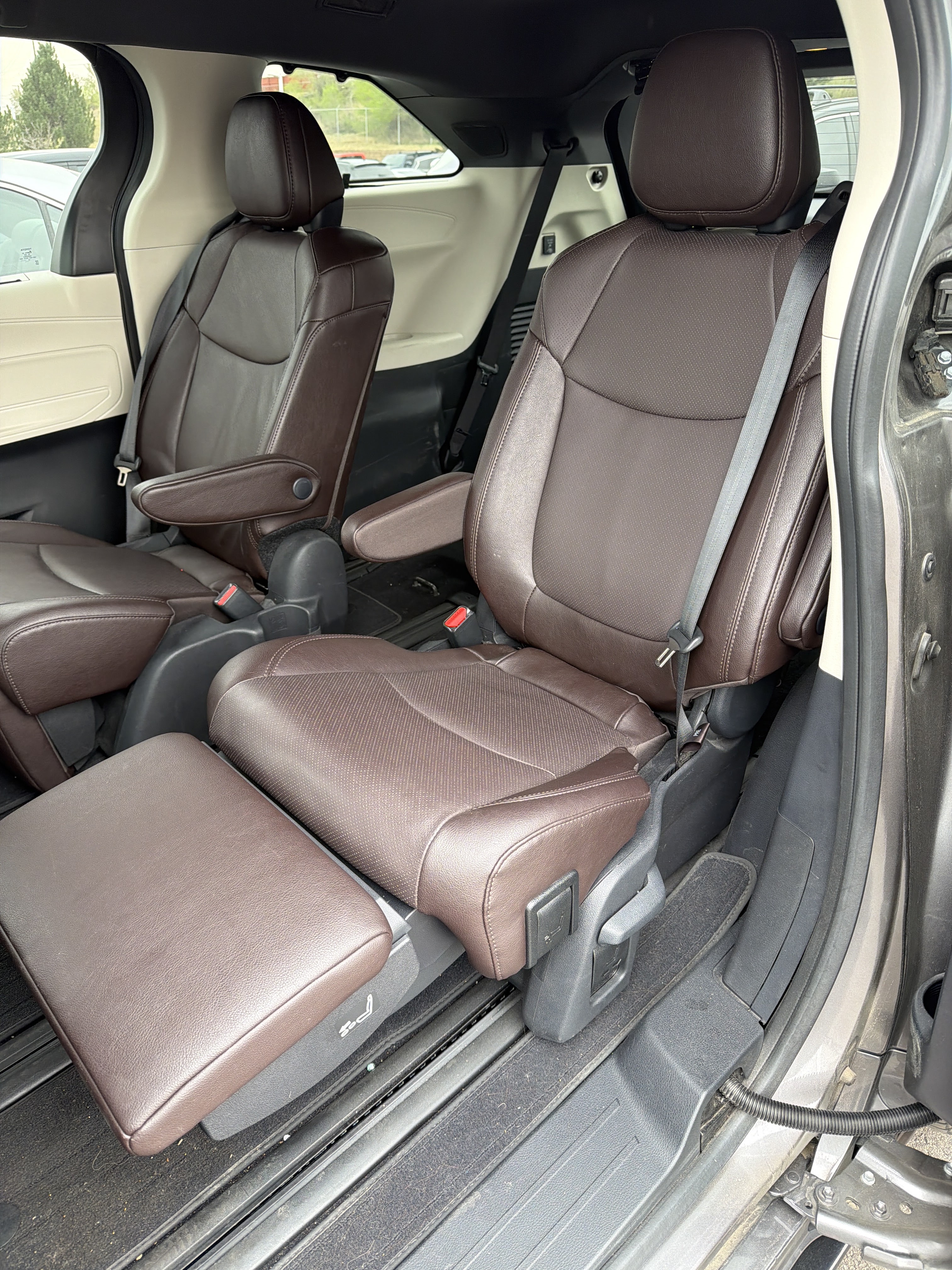 Interior view of the second row of a vehicle, showing two captain's chairs with brown leather upholstery. The chairs are equipped with armrests and adjustable footrests. The flooring is carpeted and shows some signs of wear and debris. Outside the vehicle, other cars and trees are visible through the windows.