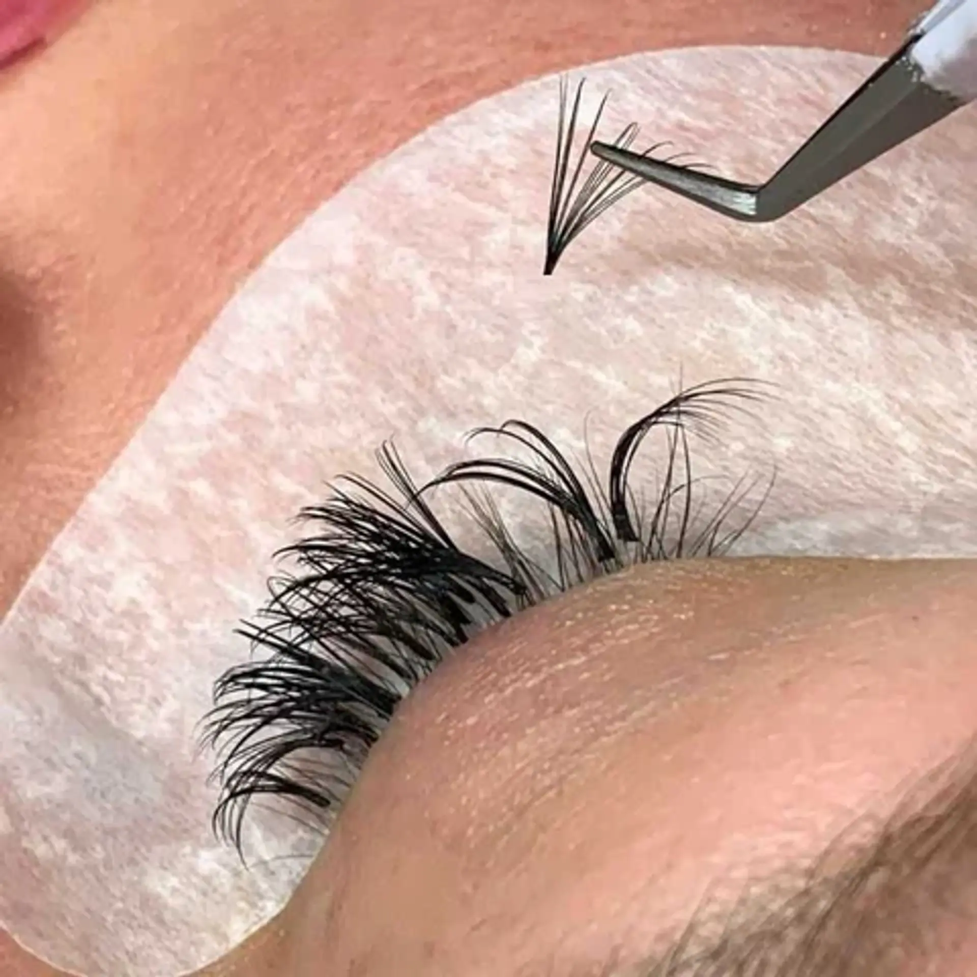 A close-up shot shows a person's eye area during a lash extension application. A white under-eye patch is placed to protect the skin. A pair of tweezers holds a fan of delicate, dark lash extensions, ready to be applied to the natural lashes. The natural lashes are already partially extended, creating a fuller look.
