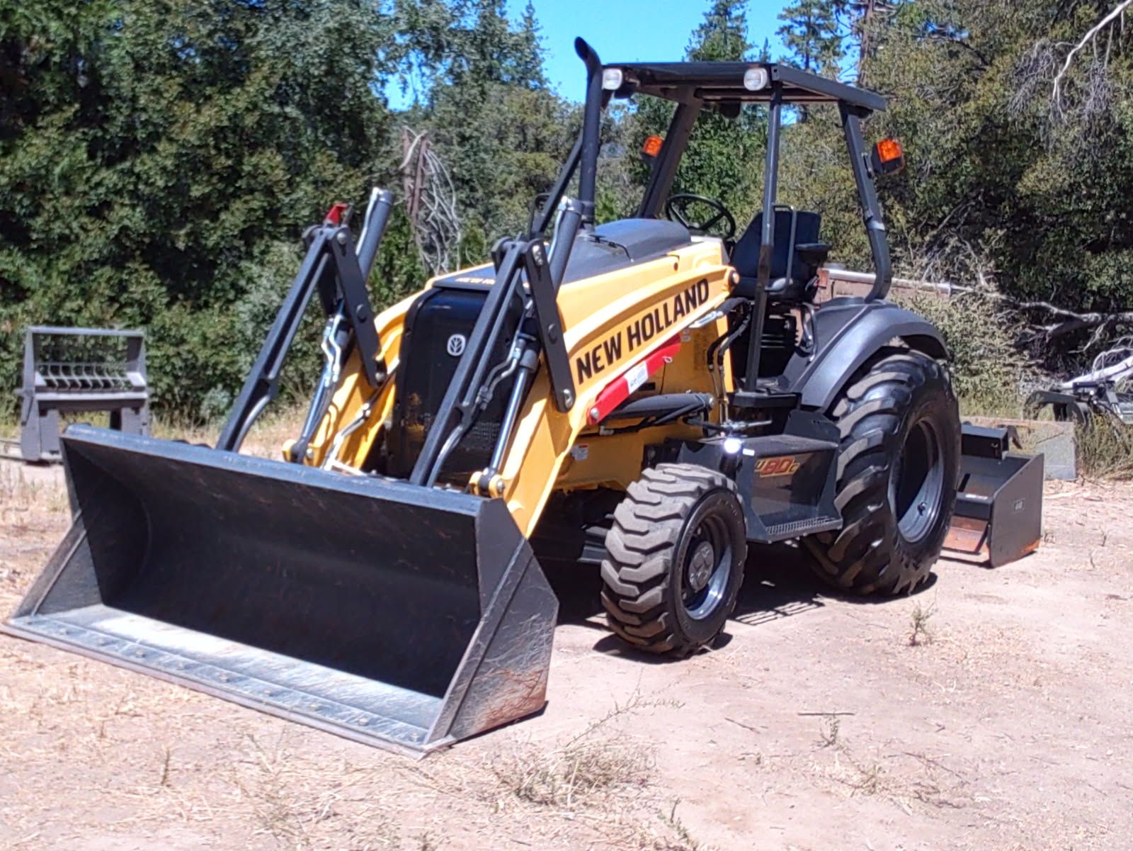 Yellow New Holland tractor-loader with a large front bucket and rear grading attachment is parked on dry ground near trees, displaying heavy-duty tires, hydraulic arms, and open operator station outdoors.