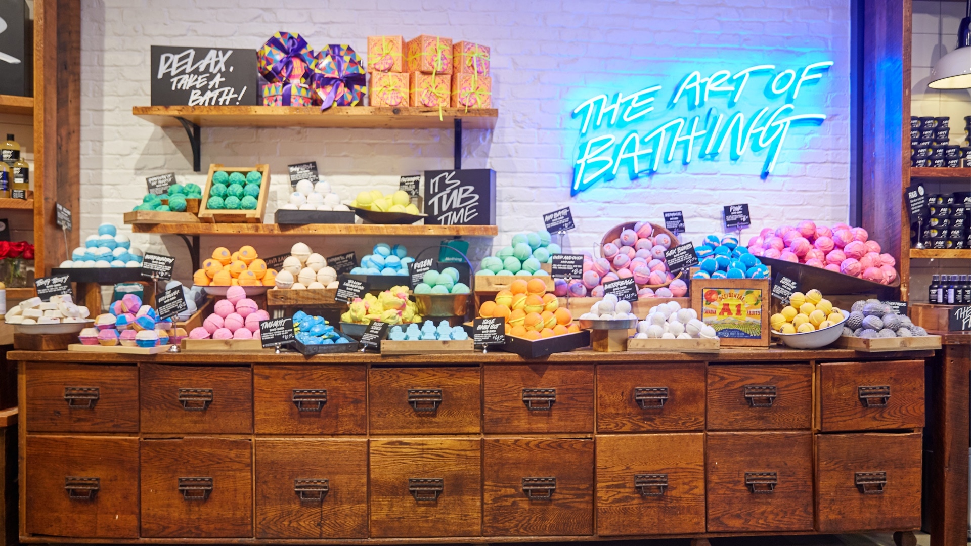 "The Art of Bathing" is written against a wall in a blue neon sign, below is a table filled with neat piles of colour-coded bath products with chalk signs that say "Relax take a Bath" and "It's Tub Time"