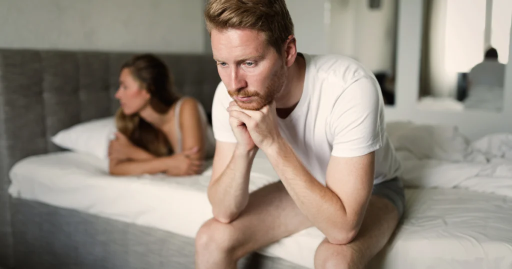 A man with red hair is sitting on the edge of a bed, looking down with his hands clasped. A woman with blonde hair is lying on the bed behind him, facing away. The room appears to be a bedroom with a gray headboard and white bedding. The lighting is soft and diffused, suggesting a natural light source.