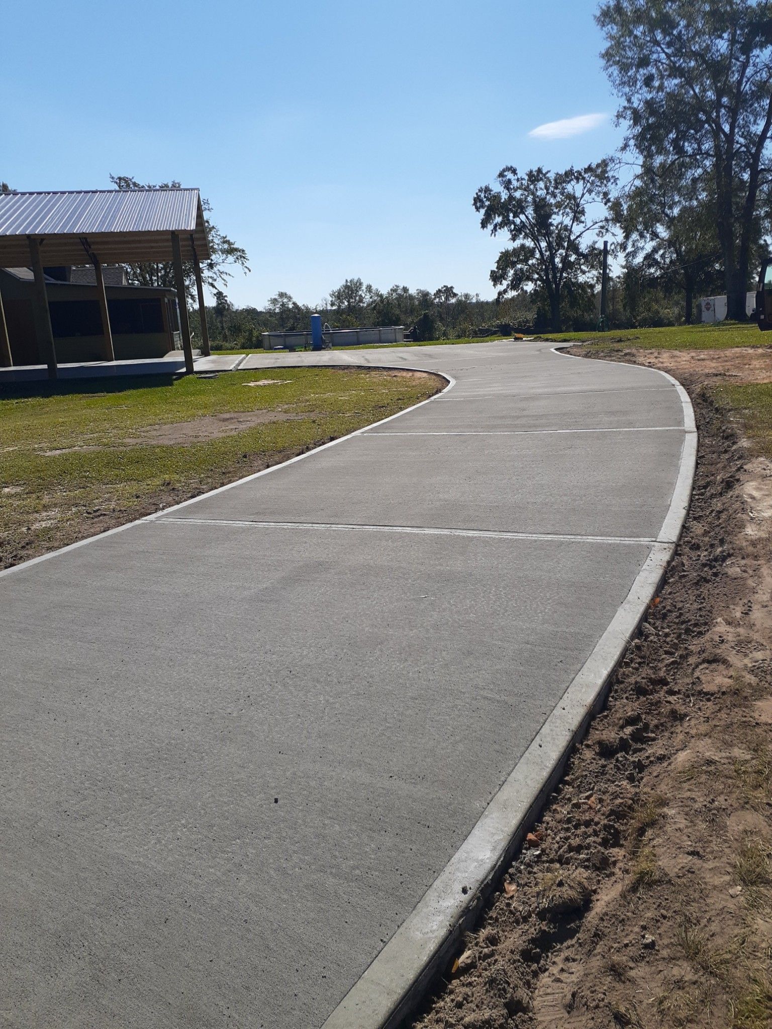 Curved concrete driveway extending through grassy rural property, smooth finished surface with expansion joints, bordered by soil edges, leading toward metal carport and trees under clear blue sky.