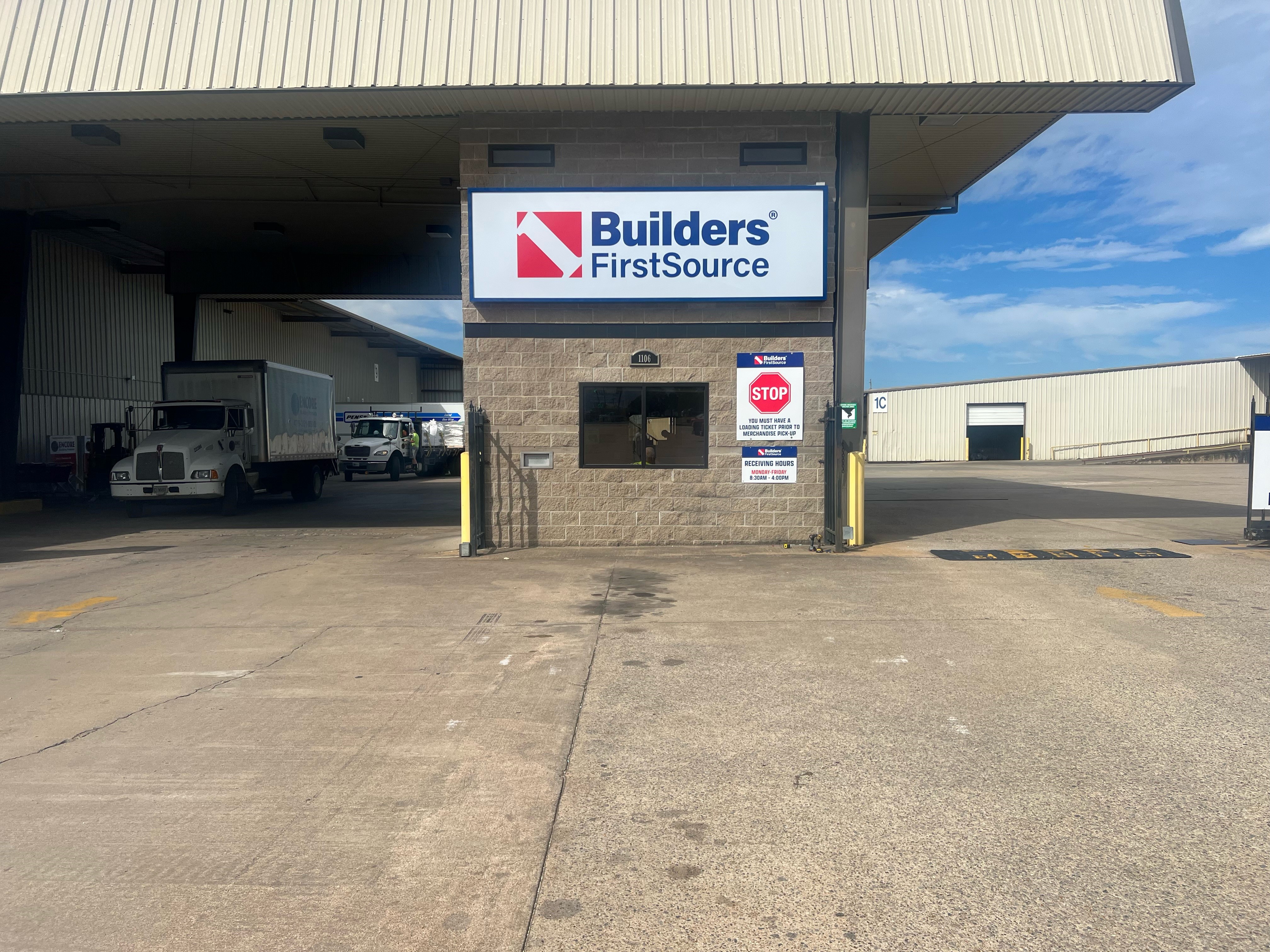 The entrance to the receiving area at a Builders FirstSource facility. A large sign with the company logo is displayed above a small window on a stone-clad building. A "STOP" sign with receiving instructions is visible, and there is a white truck parked near the entrance. The scene includes an open gate leading to the lumber yard in the background, with a clear sky above.