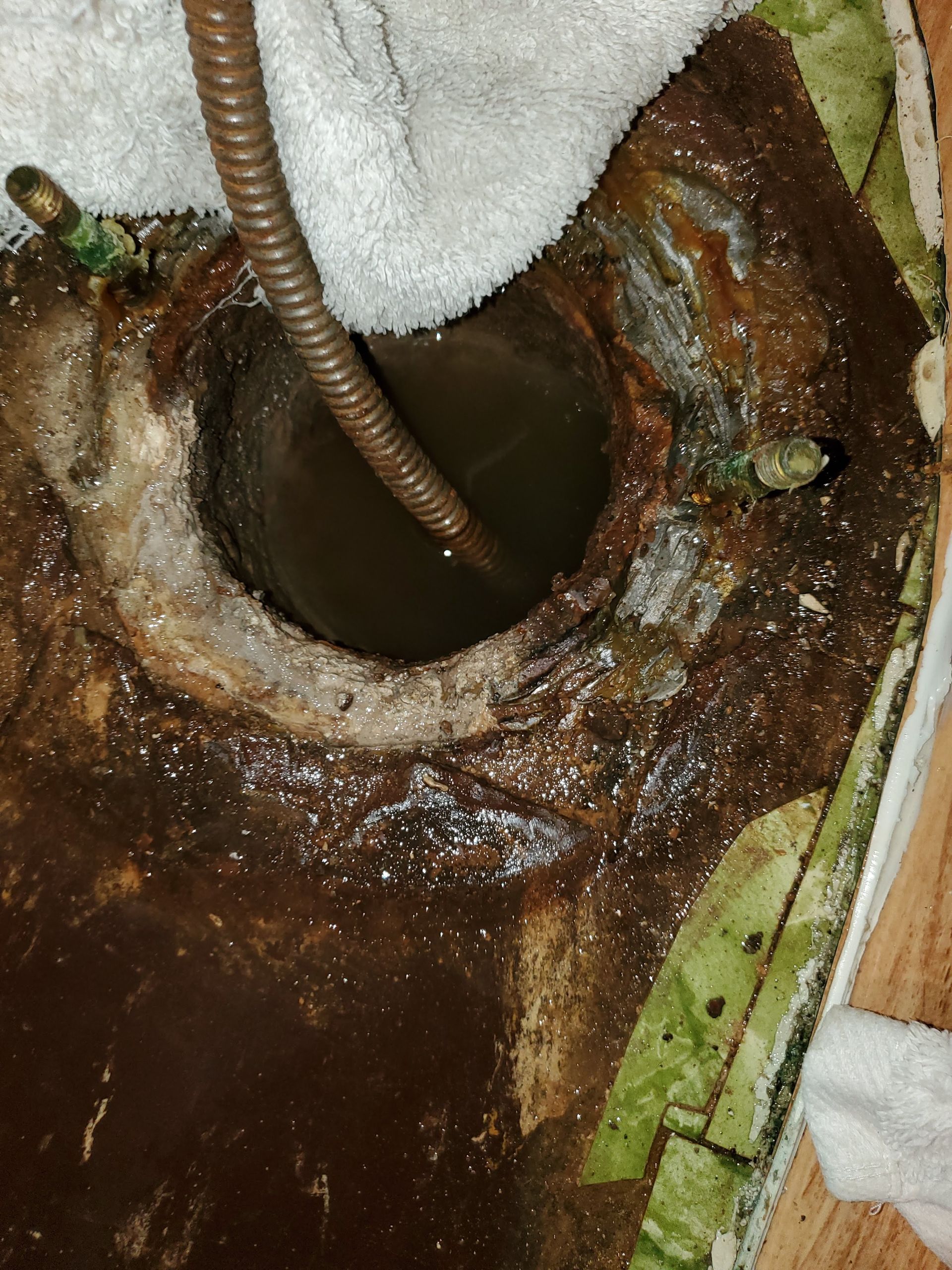 Exposed toilet drain opening with heavy rust, buildup, and dirty water visible, as a drain auger is inserted, showing severe blockage and deteriorated plumbing requiring immediate professional cleaning and repair.