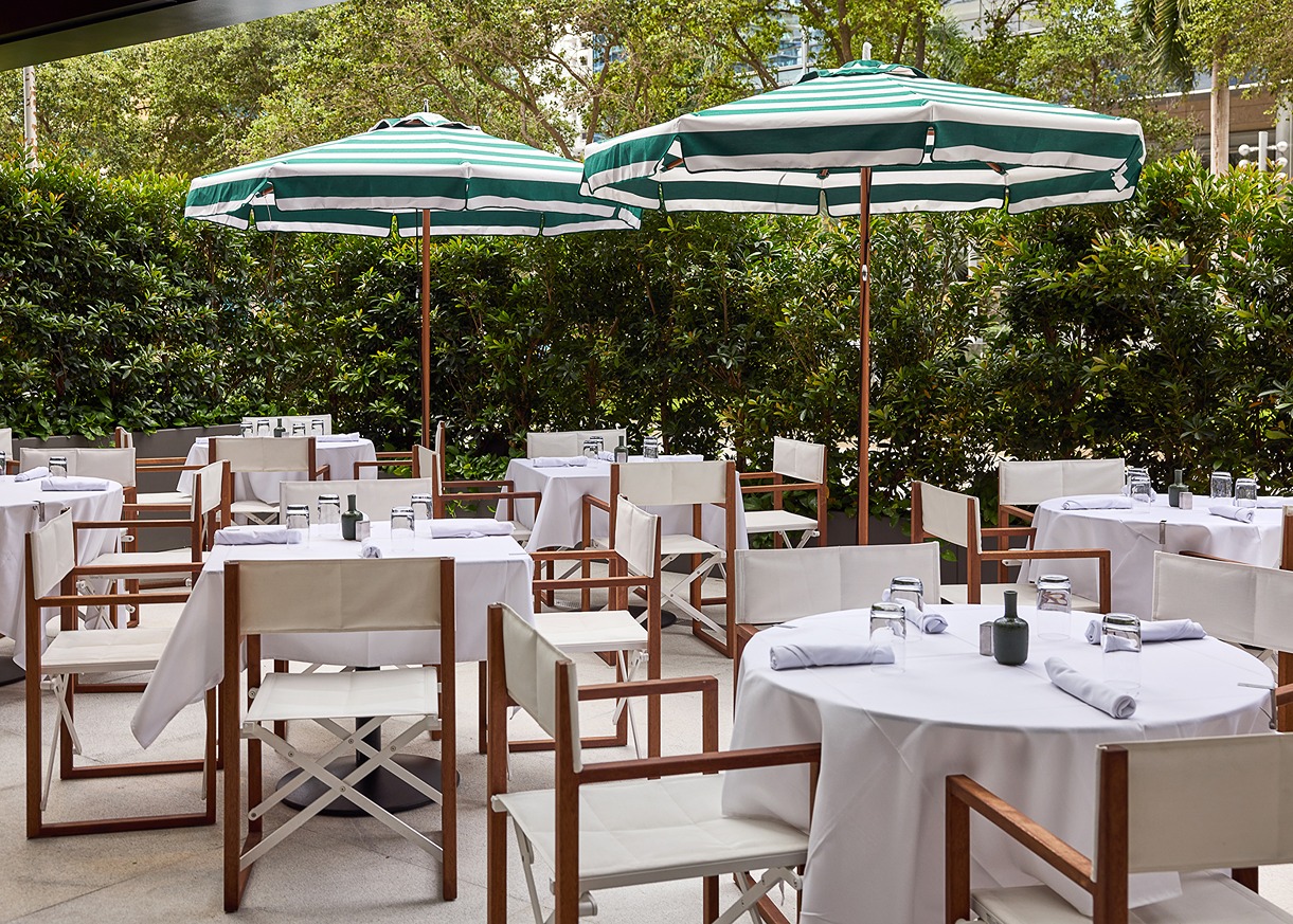 A bright al fresco dining space surrounded by lush green hedges, featuring director-style chairs and tables set for a lunch service under striped sun umbrellas.