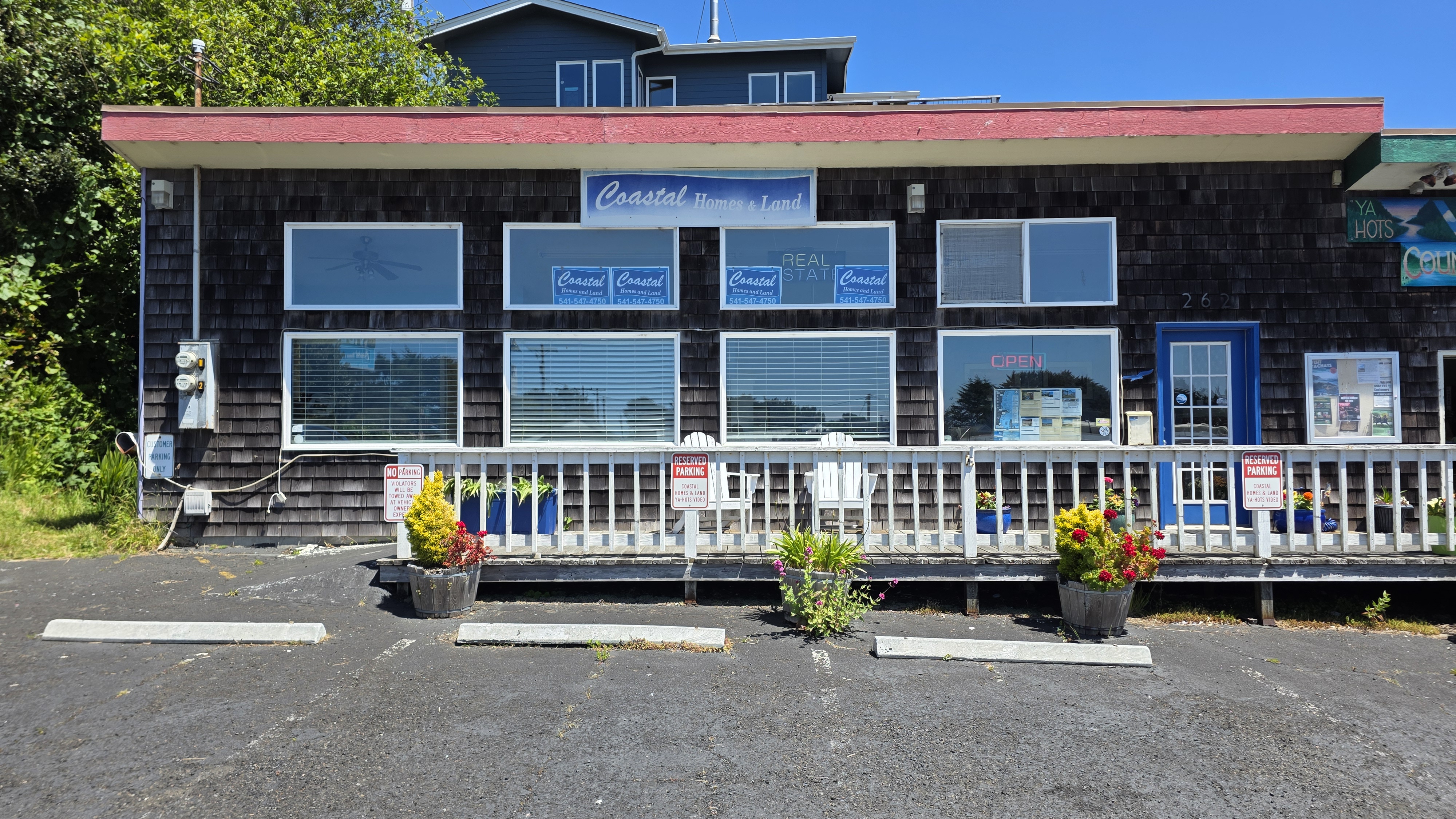 A storefront for "Coastal Homes & Land" is shown with a red overhang and dark shingle siding. There are several windows displaying "Coastal Homes and Land" signage with phone numbers. An "OPEN" sign is visible in neon red in one of the windows. A blue door is present, and a bulletin board is mounted to the wall. Potted plants with yellow and red flowers are on the porch. The storefront is located in a parking lot with concrete parking stops. The building is set against a backdrop of green trees and a clear blue sky.