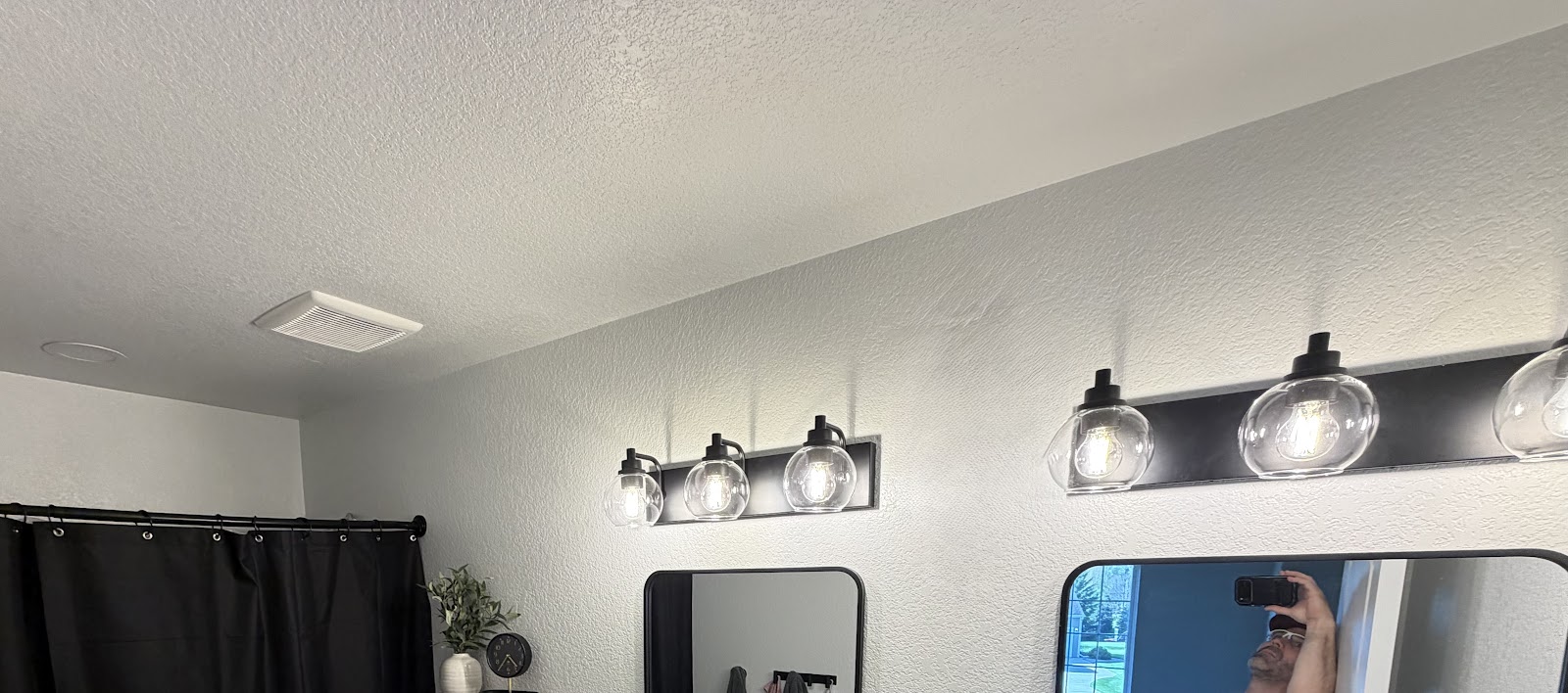 A bathroom vanity light fixture with clear glass globes is mounted above a mirror. A black shower curtain is visible on the left side of the image. The wall has a textured finish. A ventilation fan is installed in the ceiling.