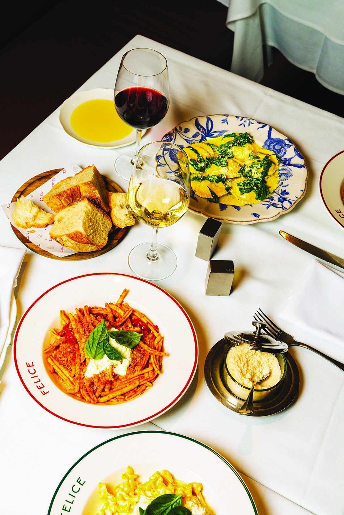 Overhead shot of fusilli with tomato and burrata, spinach ravioli, crusty Italian bread, and glasses of red and white wine. Freshly grated parmesan sits on the side.