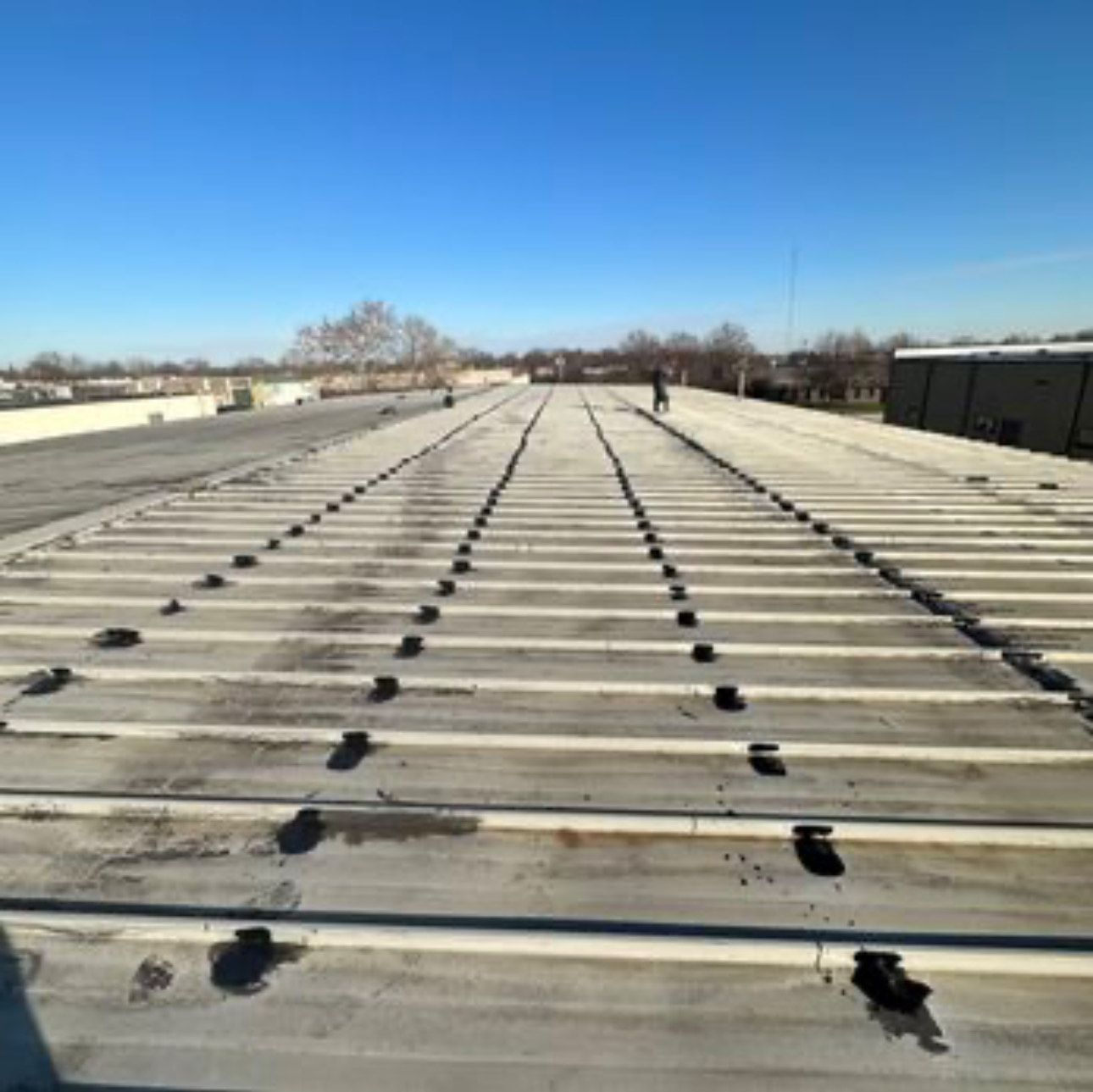 Expansive view down a long, light-colored roof featuring parallel ribs or seams. Numerous dark, regularly spaced patches or fixtures are visible along these lines. A lone figure stands distantly.
