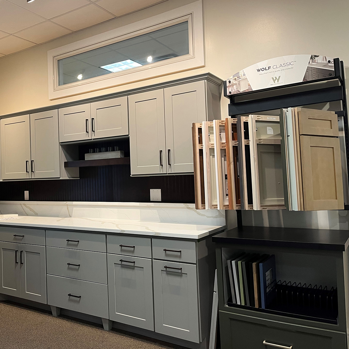 Modern kitchen display with grey shaker-style cabinets, black backsplash accent, light countertop, and a sample station showcasing cabinet doors and finishes labeled &ldquo;Wolf Classic.