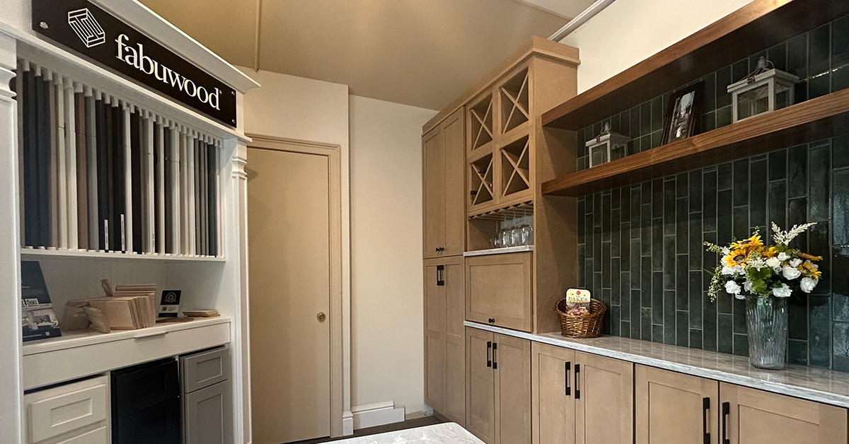 Kitchen showroom display featuring Fabuwood cabinetry with light wood finishes, a gray countertop, open shelving with decor, glass-front wine storage, and a dark green tile backsplash alongside a door and sample display wall.
