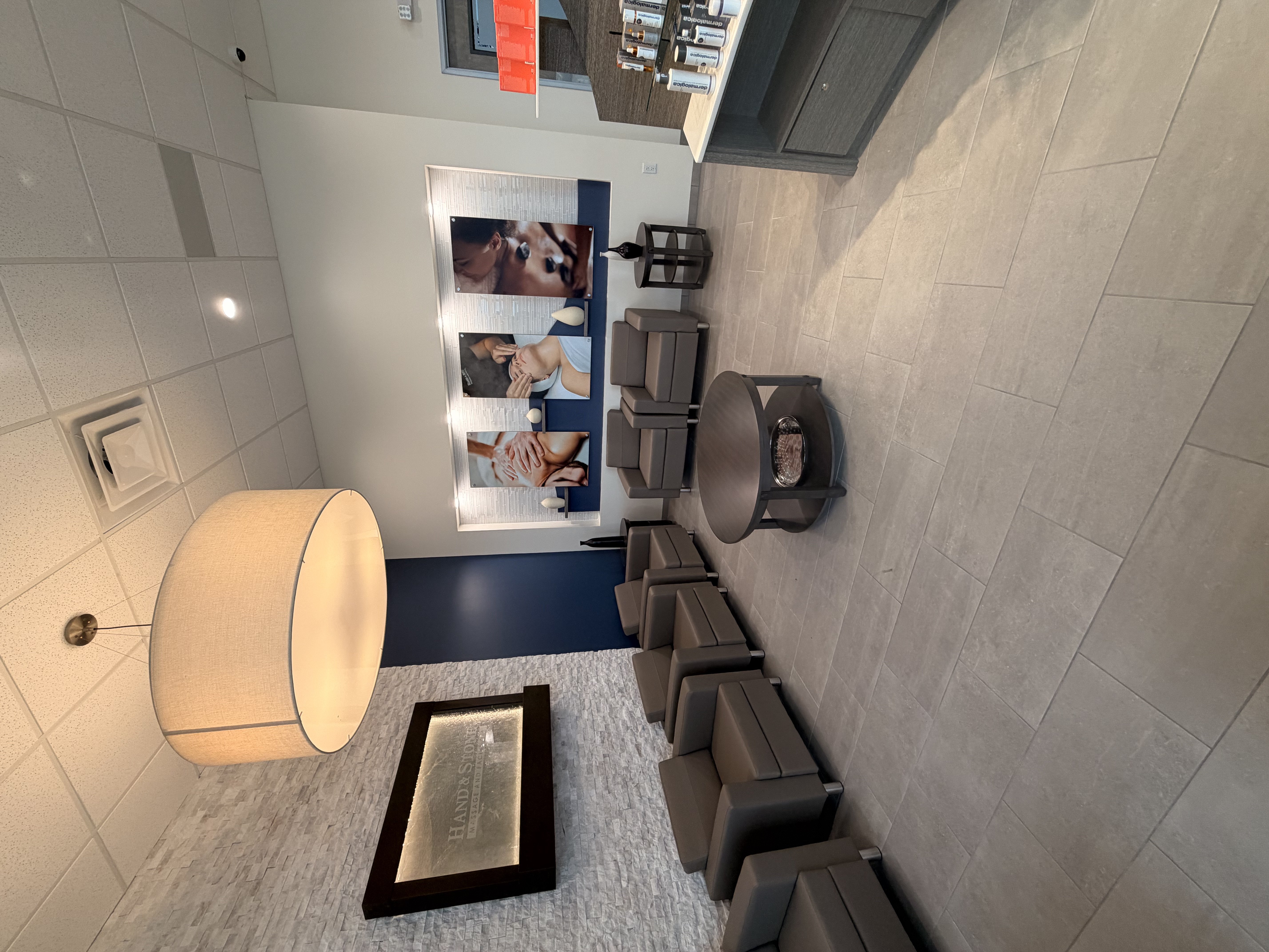 Waiting area with gray leather armchairs arranged in row. Large drum pendant light with textured shade. Stacked stone accent wall with framed artwork. Navy blue painted wall section. Gray tile flooring and small round side table.