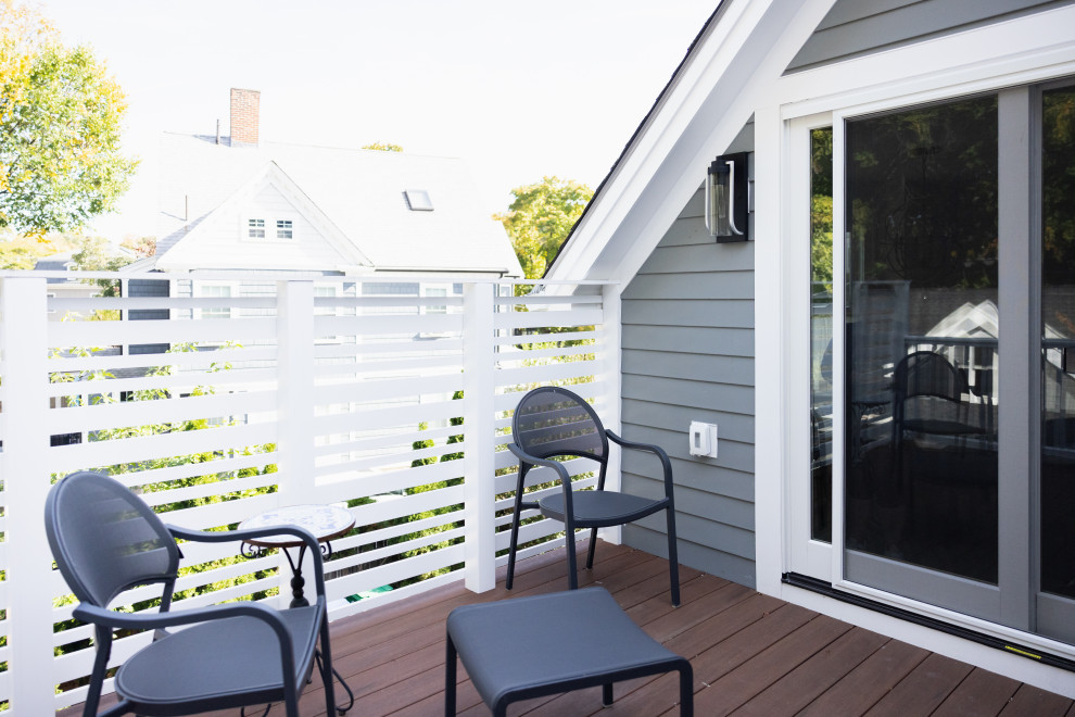 A close-up view of a modern deck with two dark gray chairs and a small square coffee table. A white slatted fence encloses the deck, with a glimpse of houses and trees in the background. A sliding glass door leads into the building.